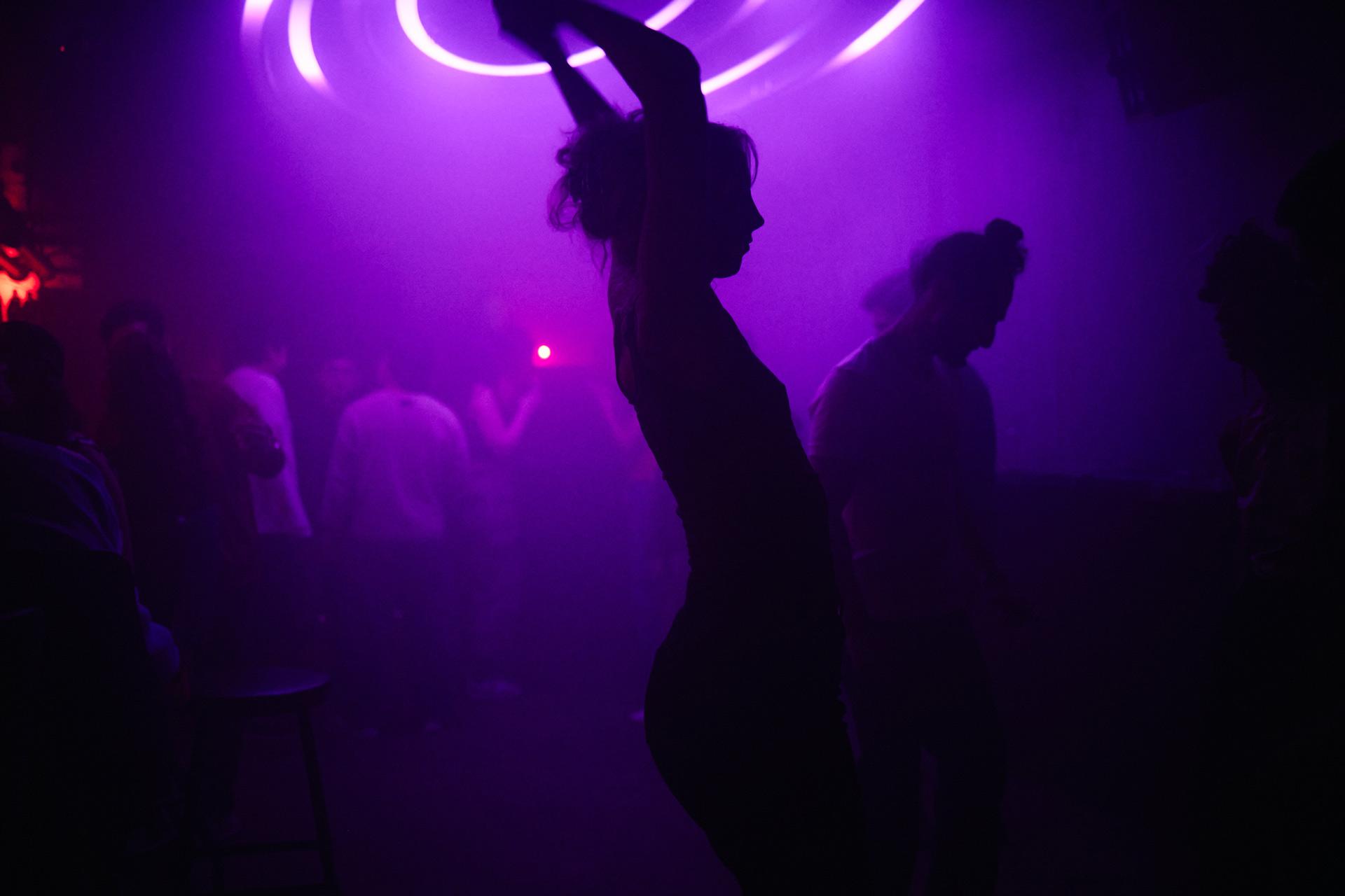 A silhouette of a person dancing with arms raised in a dark club, illuminated by purple neon lights, while other people mingle and dance in the smoky background.