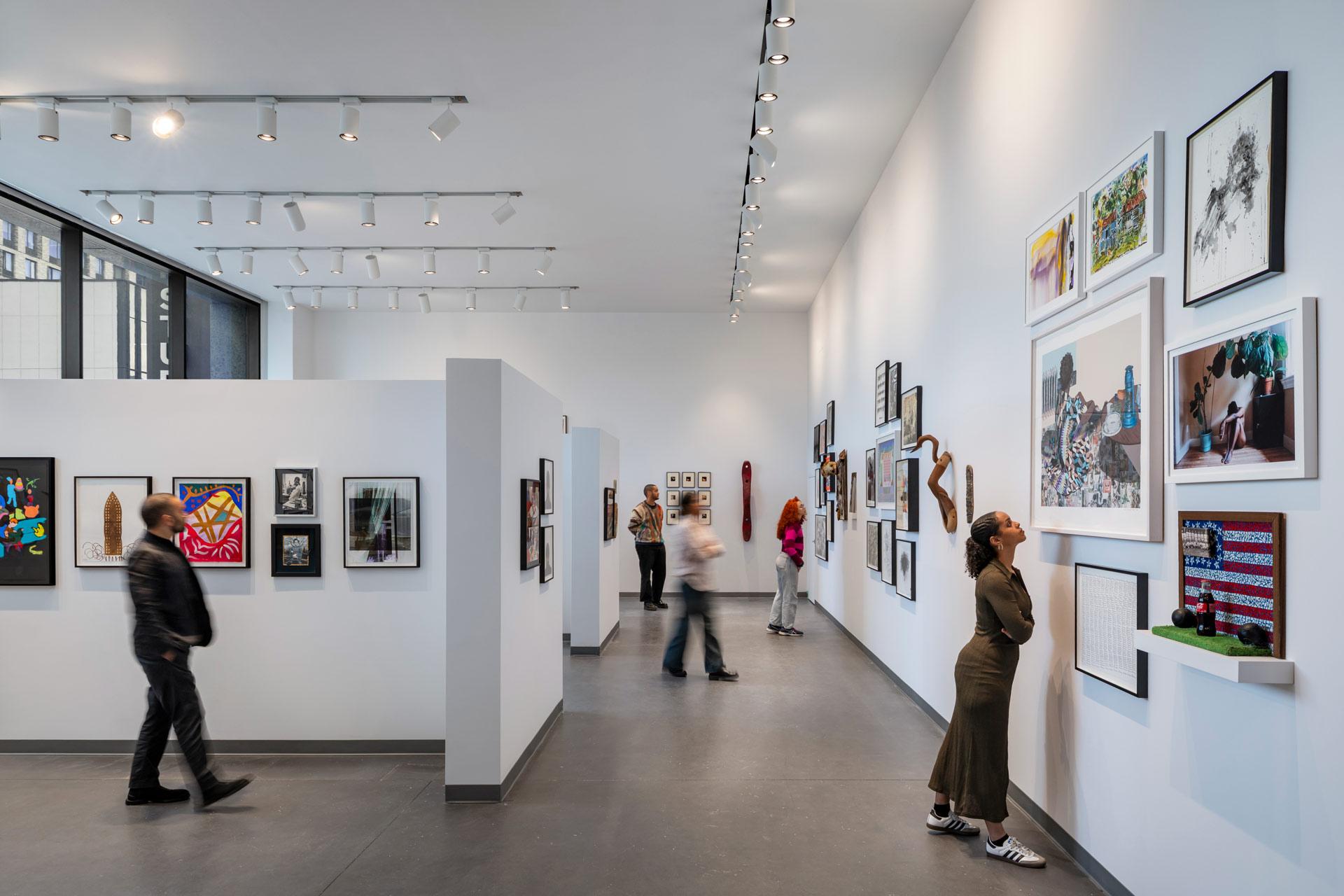 A modern art gallery with white walls displays various artworks. Several people walk, stand, and observe the pieces. Large windows let in natural light.