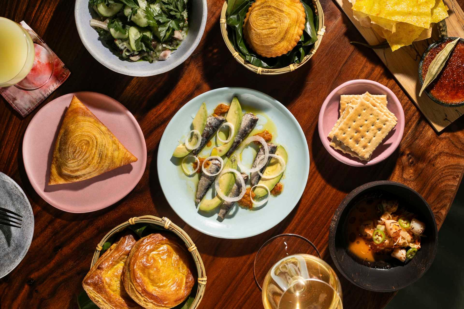 A tabletop with assorted dishes, including smoked fish with avocado and onions, leafy greens, crackers, puff pastries, a golden pastry triangle, kimchi, drinks, and crispy chips on wooden and ceramic plates.