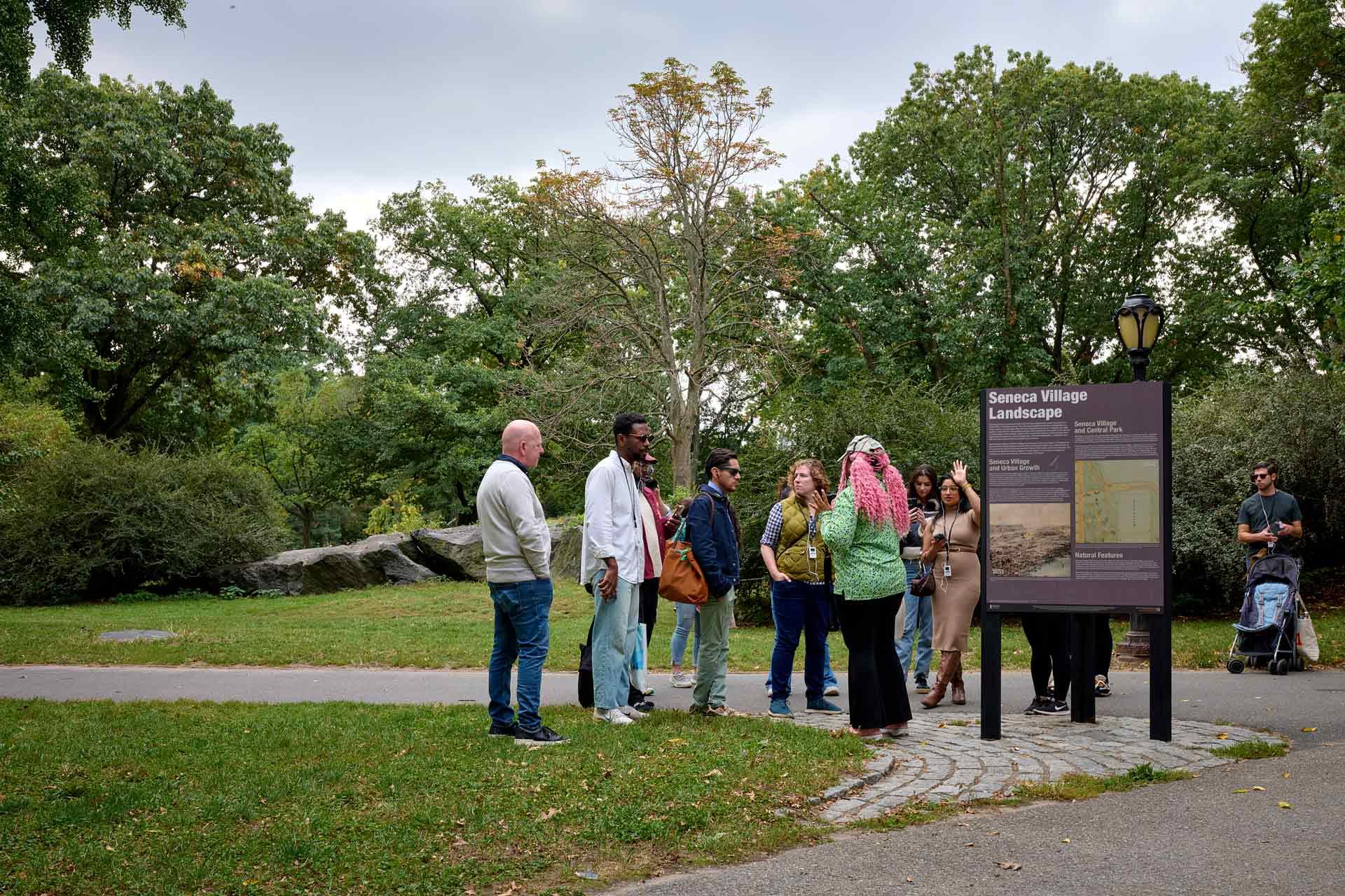 A diverse group of people stand near an informational sign about Seneca Village Landscape in a park surrounded by green trees and grass. Some listen attentively while others look around or chat.