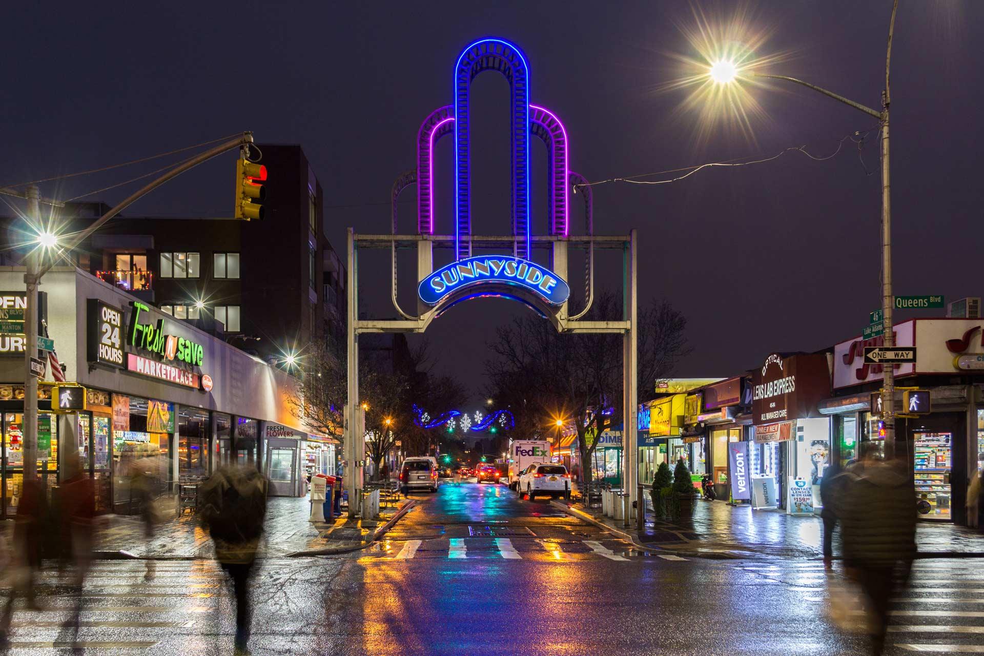 A nighttime street scene in Sunnyside, Queens, New York, featuring a glowing neon "Sunnyside" sign and a colorful art deco building in the background. Pedestrians cross the wet street lined with shops and lights.