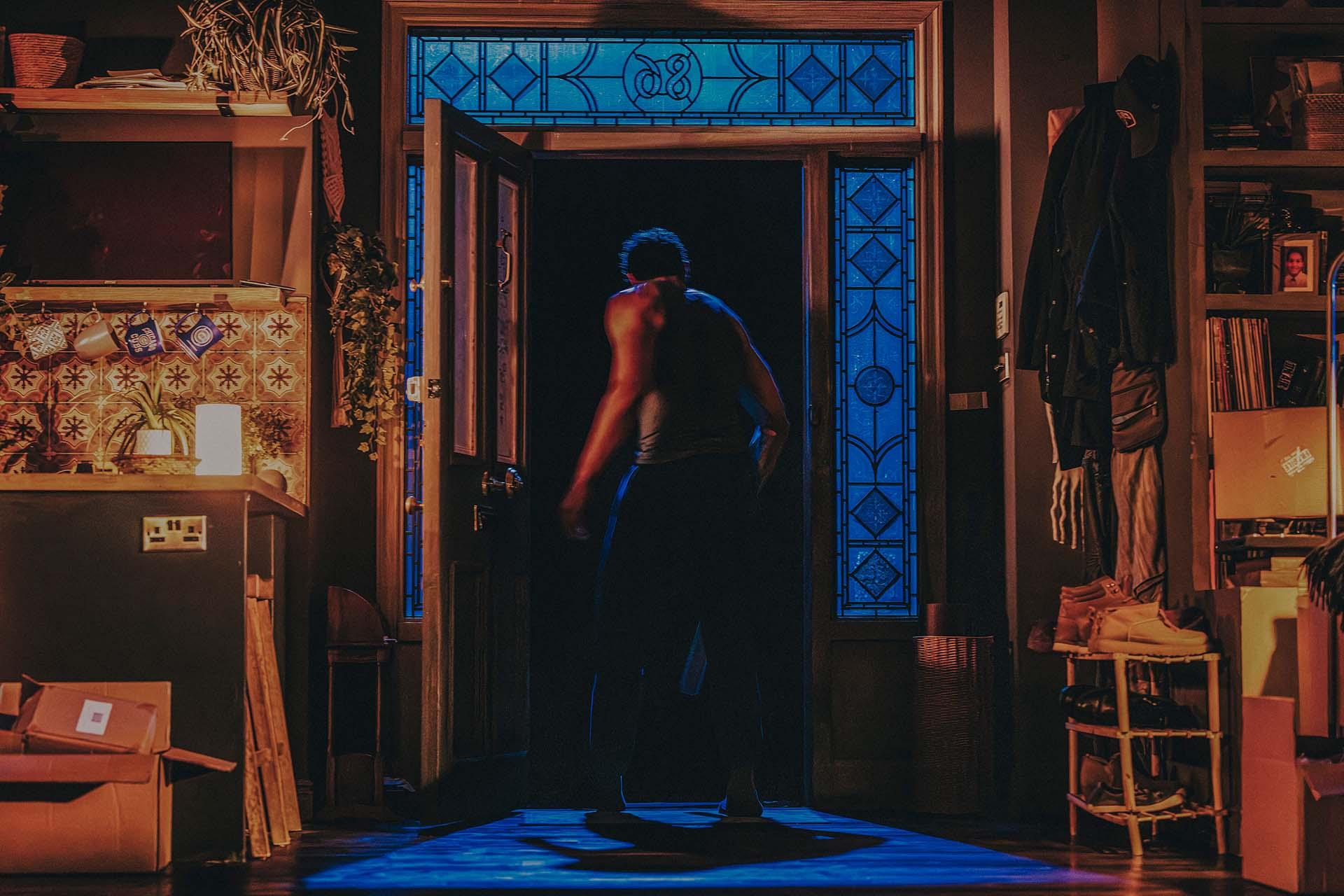 A person stands in a dimly lit room facing an open door with blue stained glass, casting blue light inside. Shelves, coats, and boxes fill the room, creating a cozy, lived-in atmosphere.