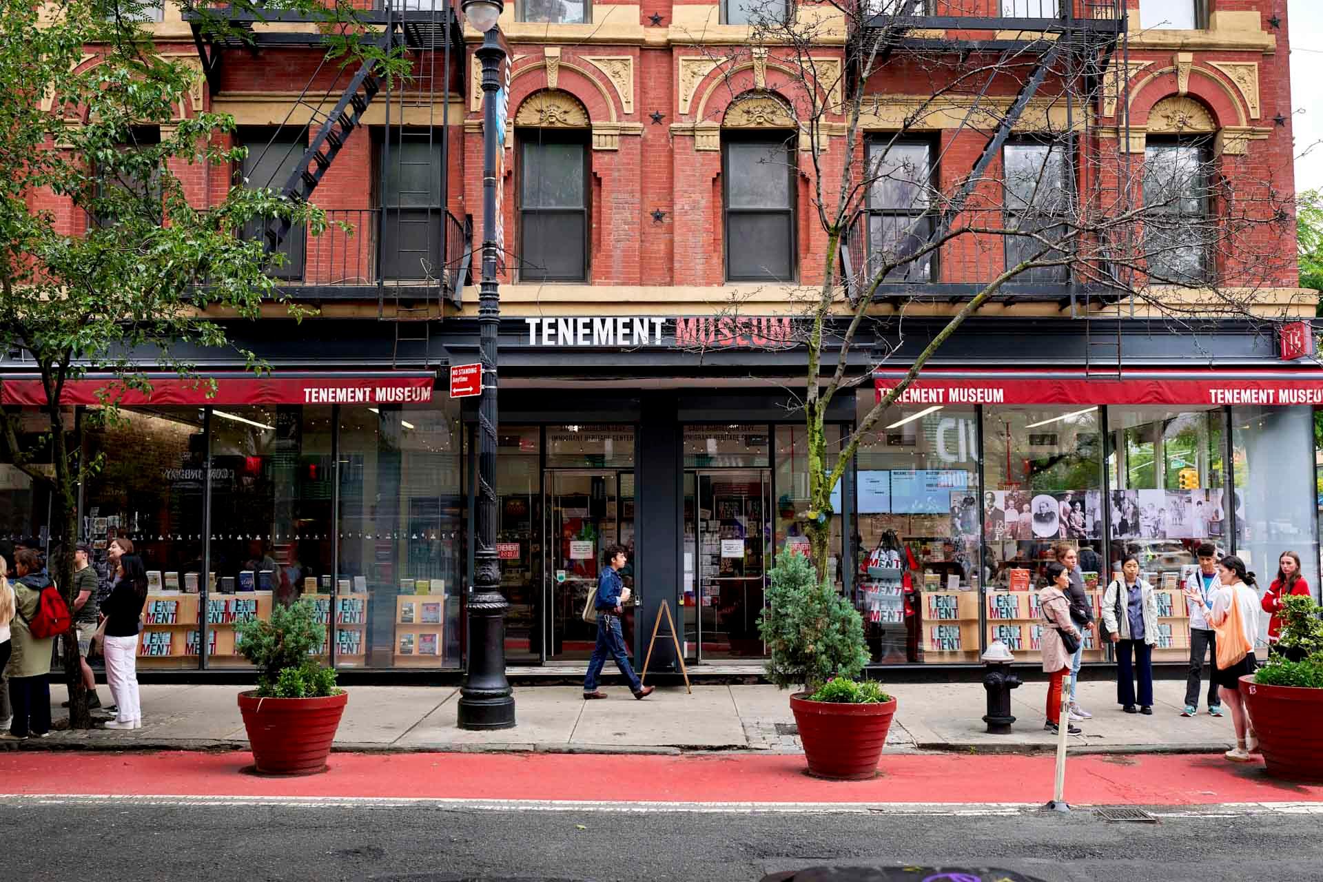 Street view of the Tenement Museum, a red-brick building with large glass windows, people standing and walking on the sidewalk, and potted plants lining the street.