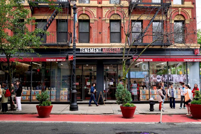 Street view of the Tenement Museum, a red-brick building with large glass windows, people standing and walking on the sidewalk, and potted plants lining the street.