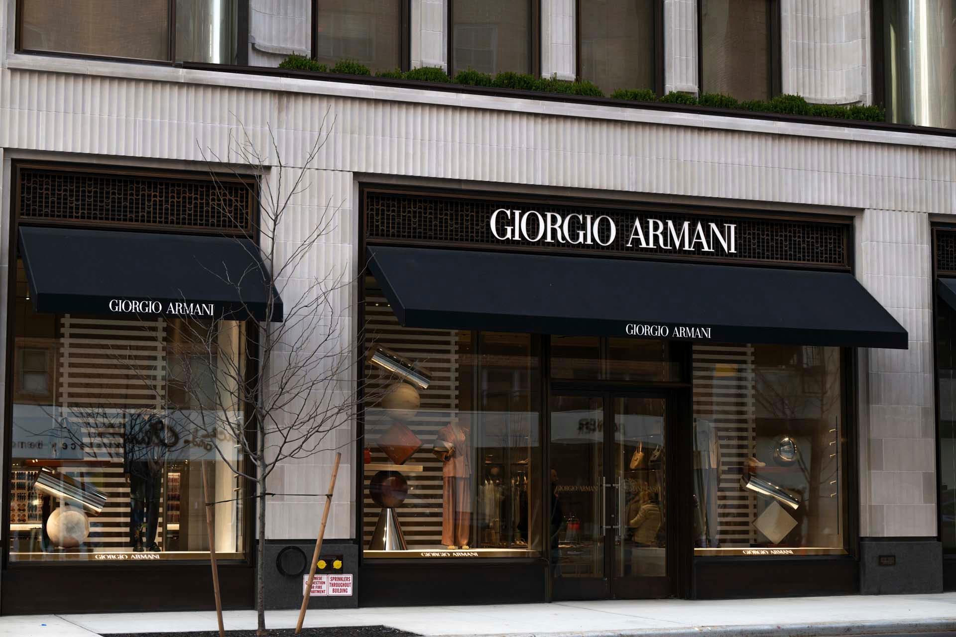 Storefront of Giorgio Armani with large windows and black awnings displaying the brand's name. Reflections of the street and interior decor are visible through the windows.