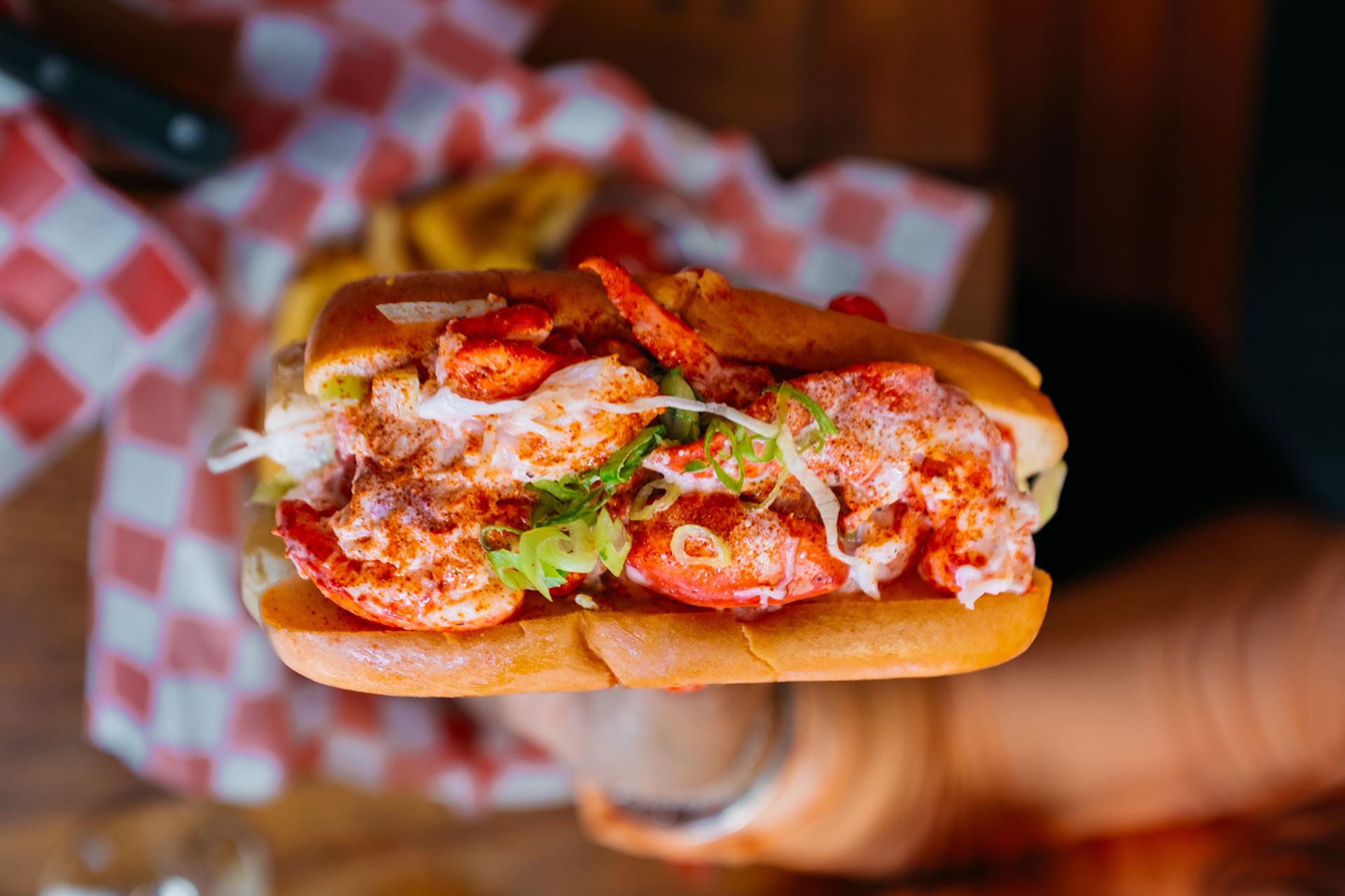 A close-up of a lobster roll with chunks of lobster meat, lettuce, and seasoning in a toasted bun, held by a hand above a checkered food paper background.