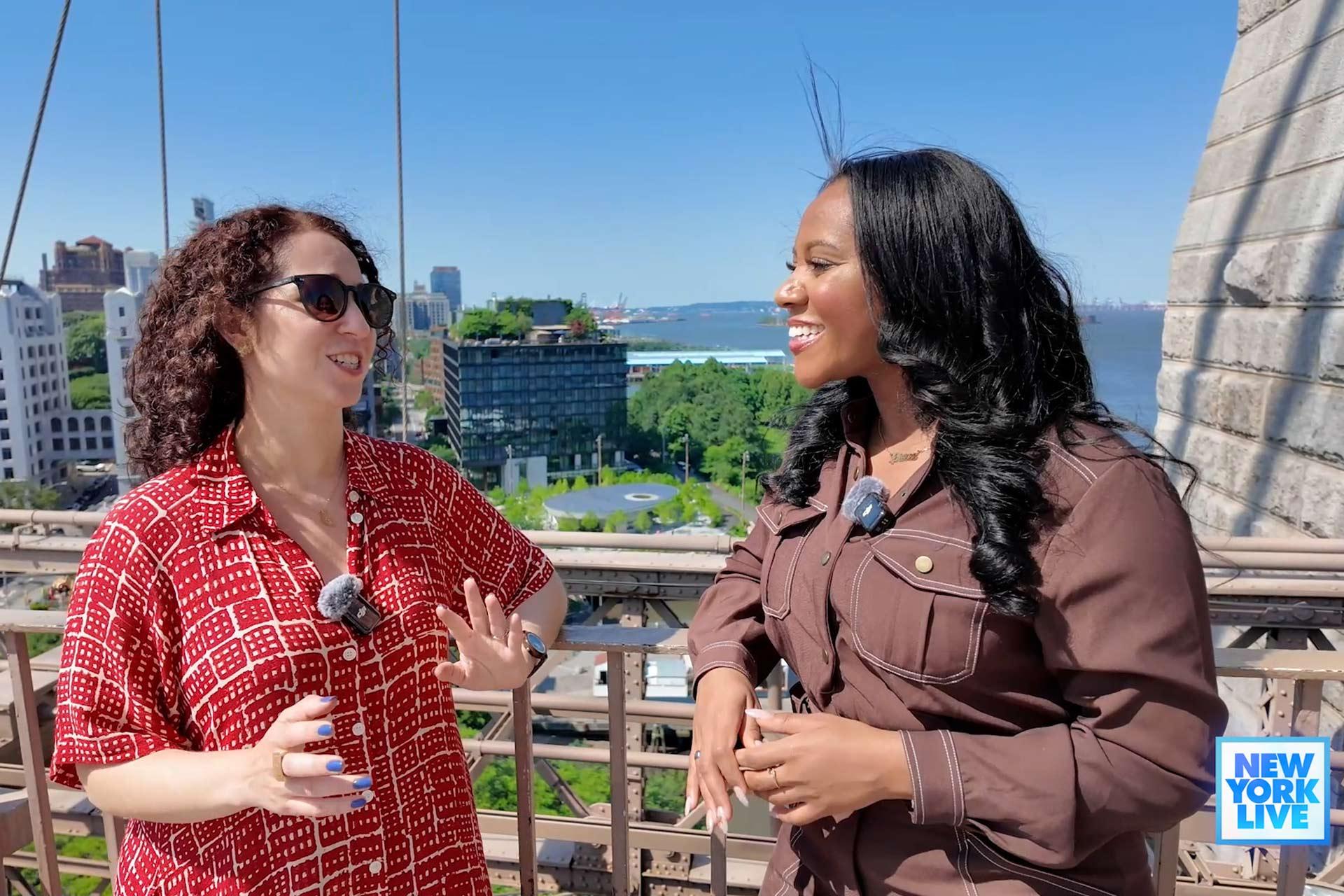 Two women stand outdoors on a sunny day, smiling and talking, with city buildings and greenery in the background. Both wear clip-on microphones. A "New York Live" logo is visible in the lower right corner.