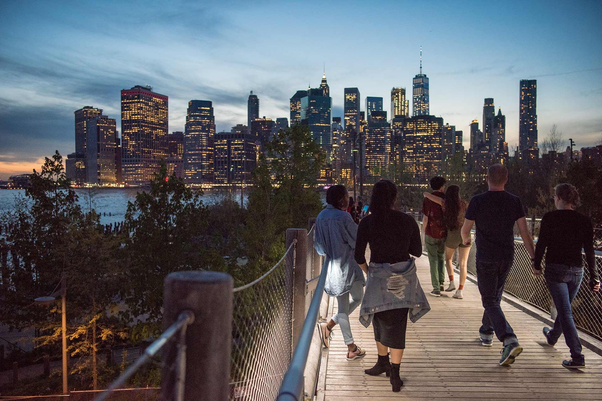 People walk along a wooden path with trees on either side, overlooking a river and the illuminated New York City skyline at dusk. The city lights reflect on the water under a blue evening sky.