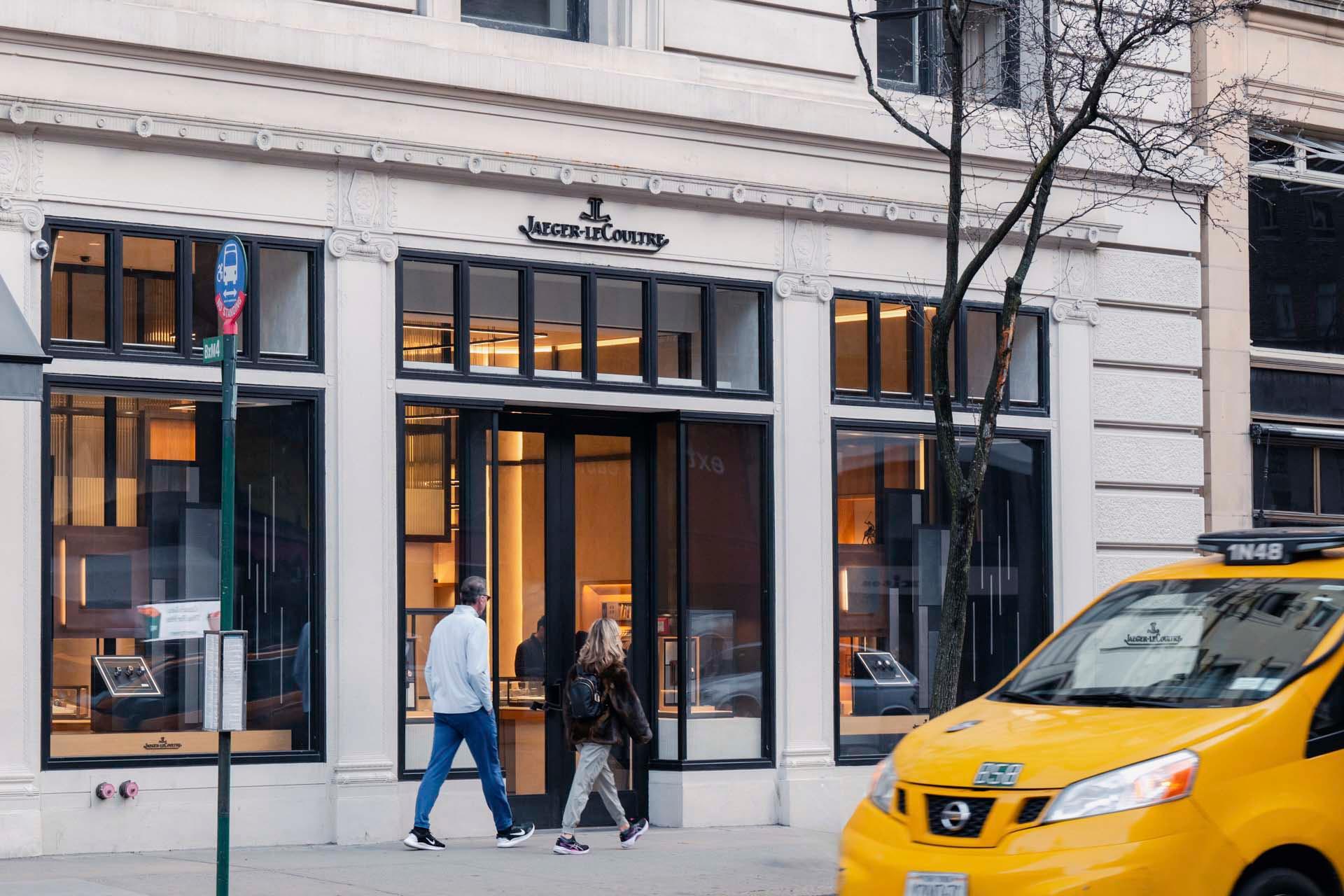 A man and a woman walk past a Jaeger-LeCoultre store on a city street. The facade of the store is elegant with large windows displaying products. A yellow taxi is passing by in the foreground.