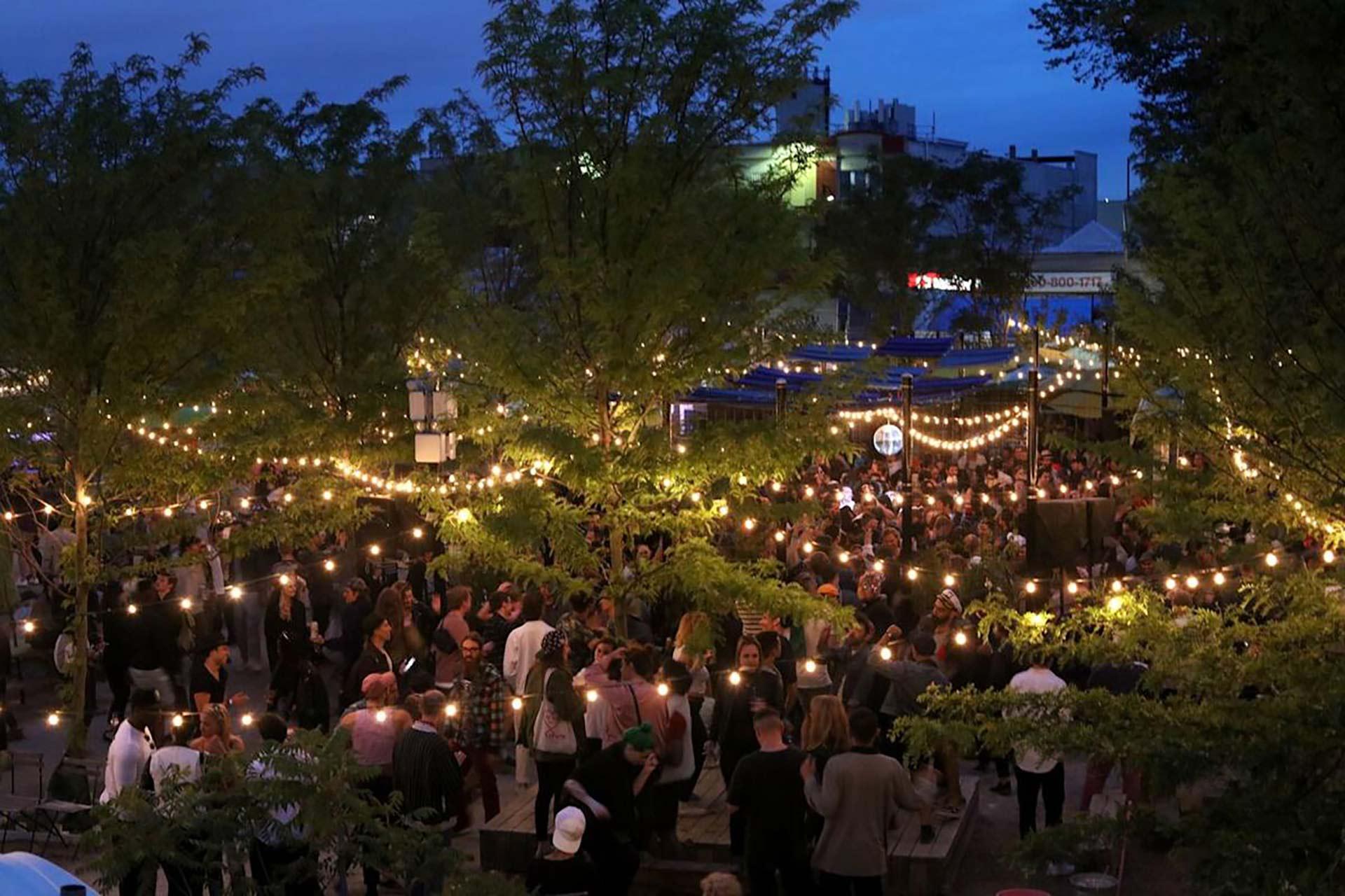 A large, diverse crowd of people gathered in an outdoor courtyard illuminated by strings of hanging lights at dusk.