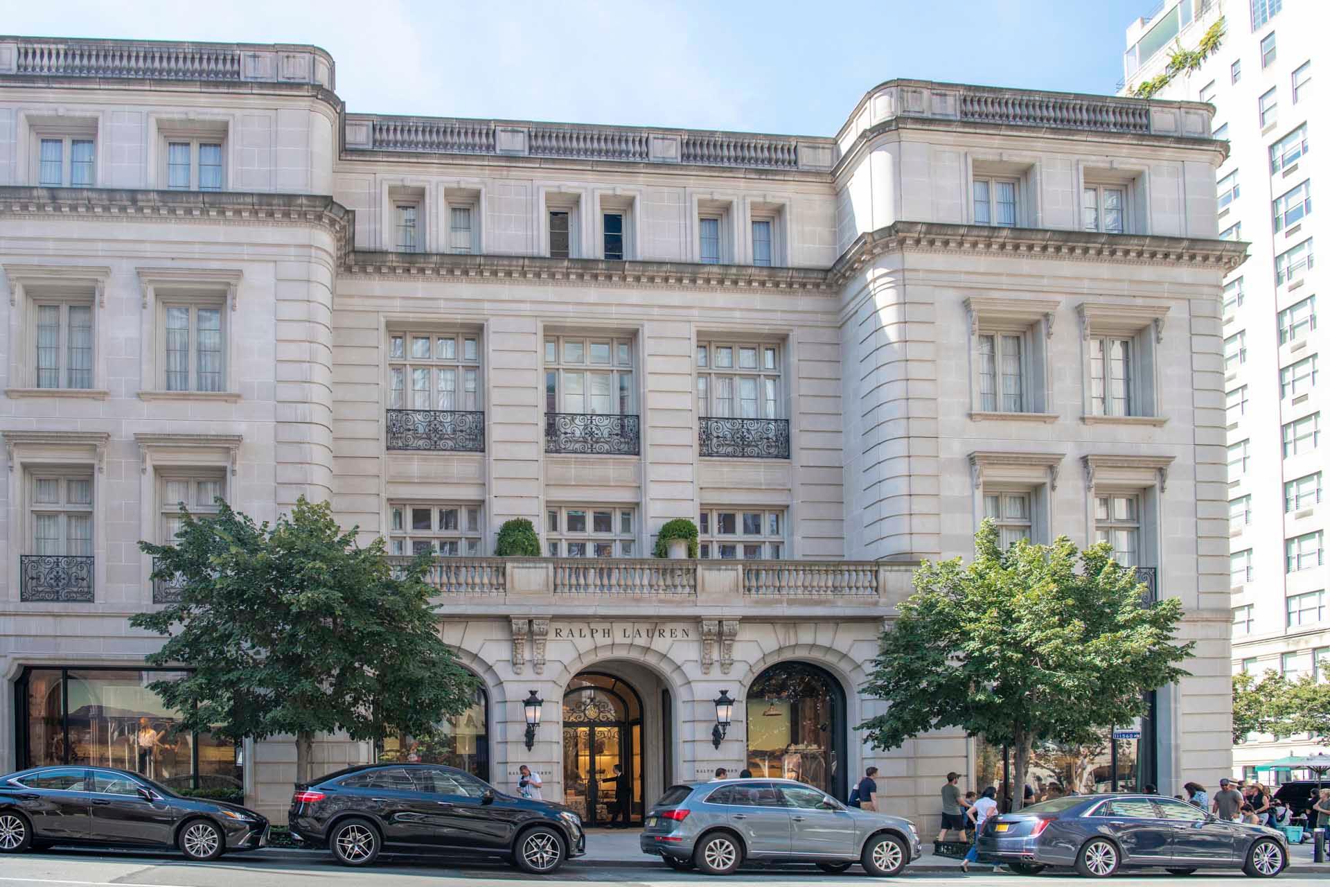 Elegant, multistory building with classical architecture, featuring large windows and ornate balconies. Trees and cars line the street below. Signs with text are above entrance doors, and people walk along the sidewalk.