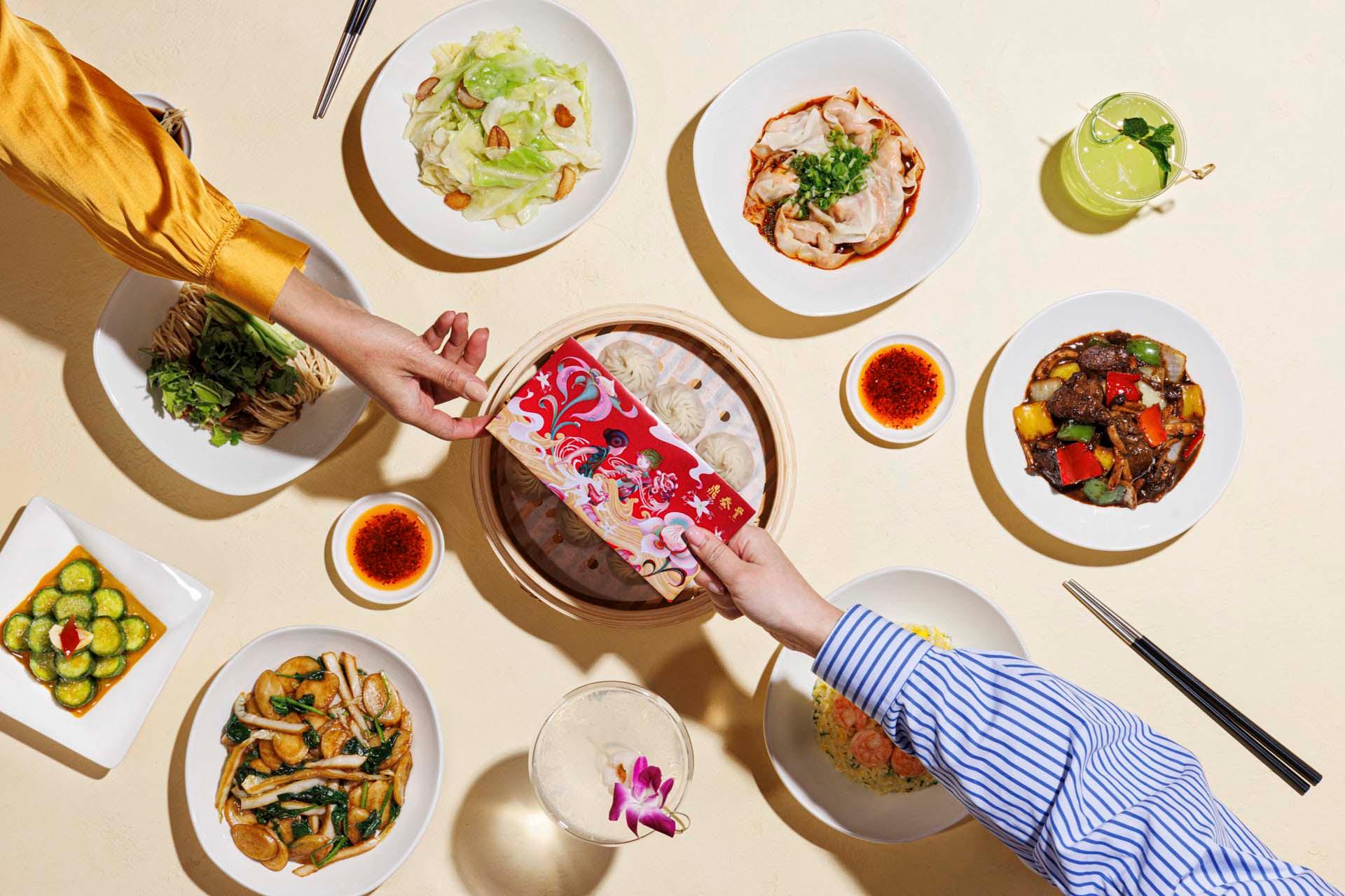 Two people sharing a colorful meal at a round table. The table is set with various Chinese dishes, including dumplings, noodles, and vegetables. Chopsticks and dips are also visible. A vibrant red envelope is being exchanged.