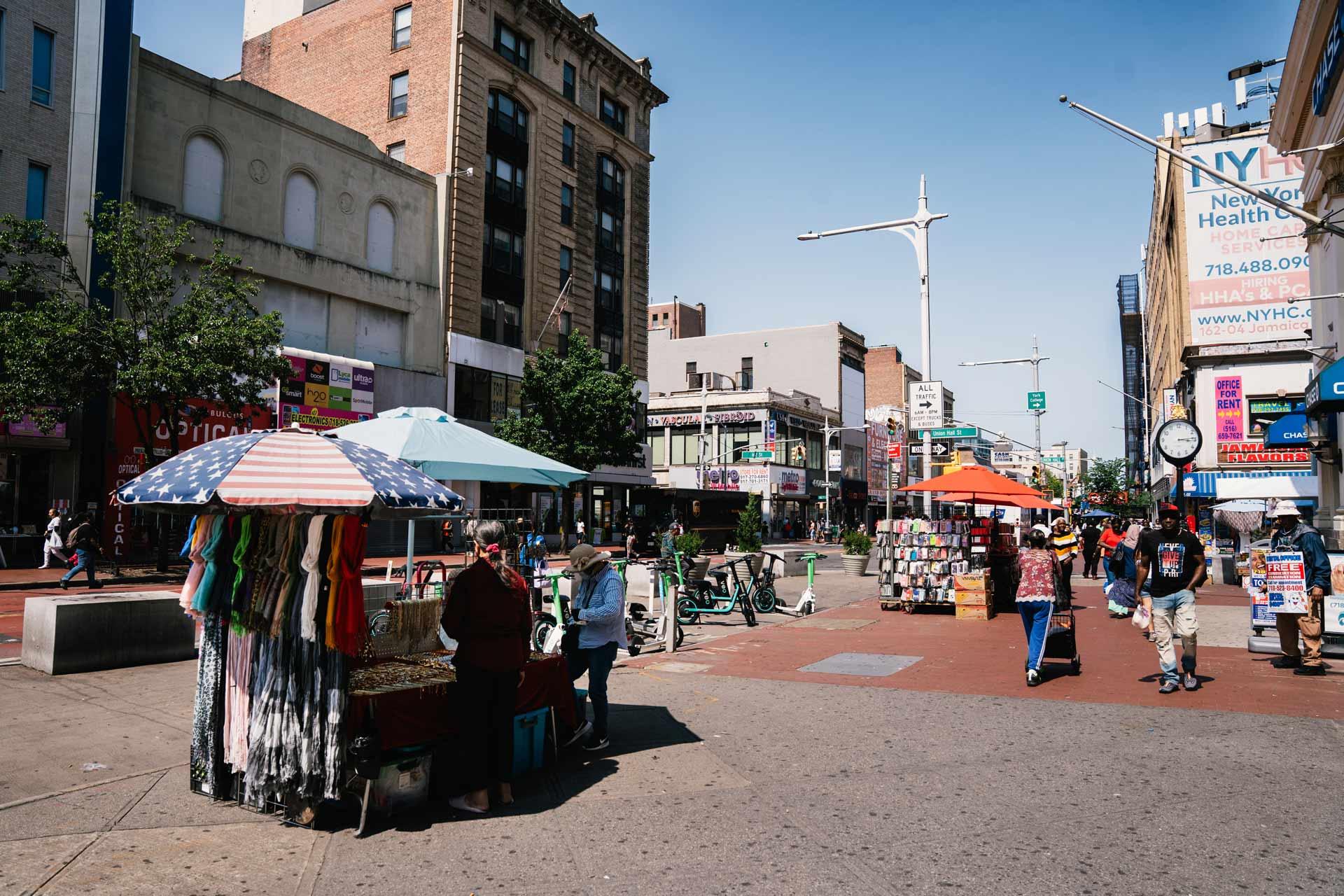 A Jamaica Avenue scene with people walking, vendor stalls with umbrellas selling goods, and tall buildings lining both sides of the street under a clear blue sky.