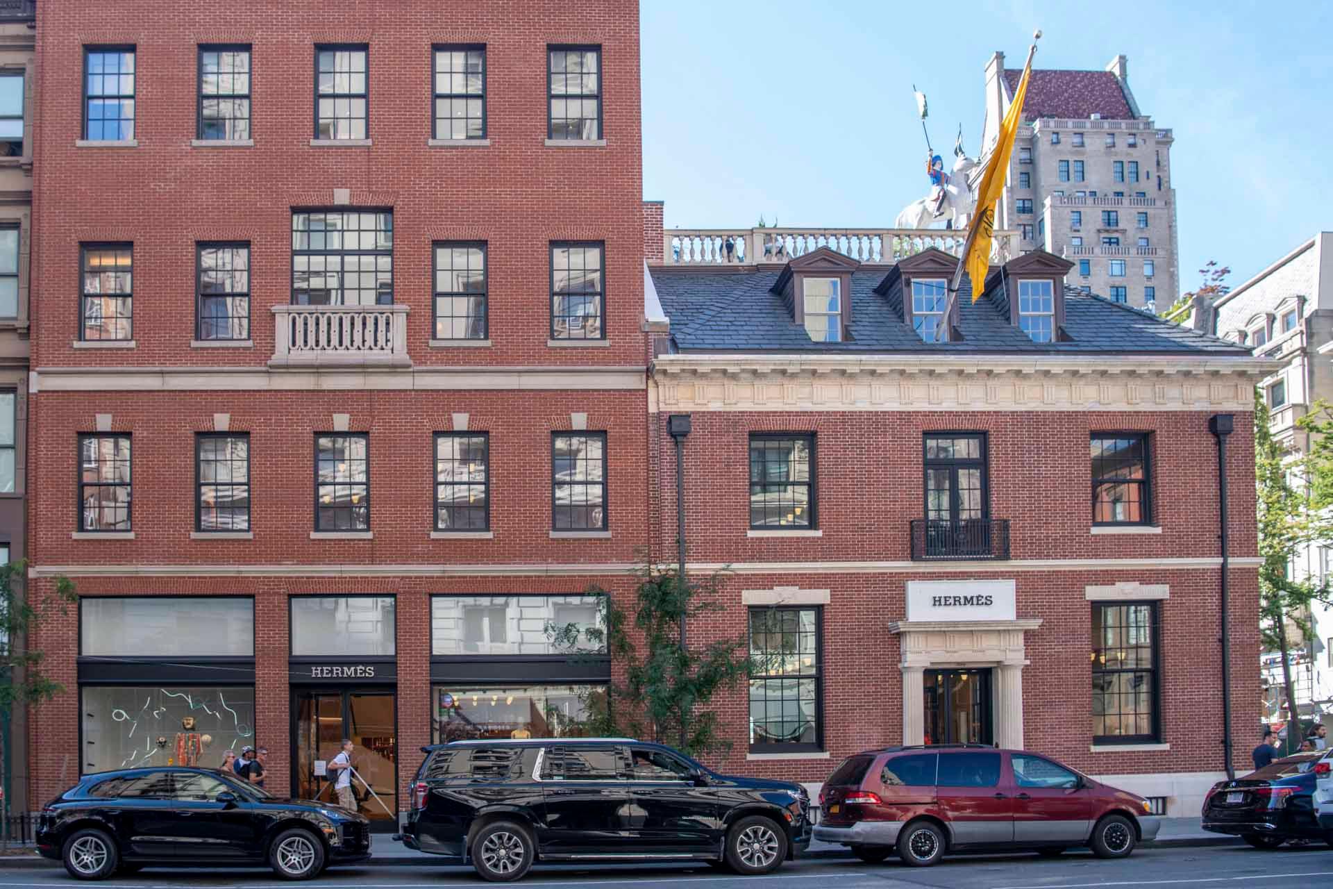 A red-brick Hermès store on a city street. The building has a classic design with two stories and large windows displaying products. Several parked cars line the street in front, and a flag with the Hermès logo hangs above the entrance.