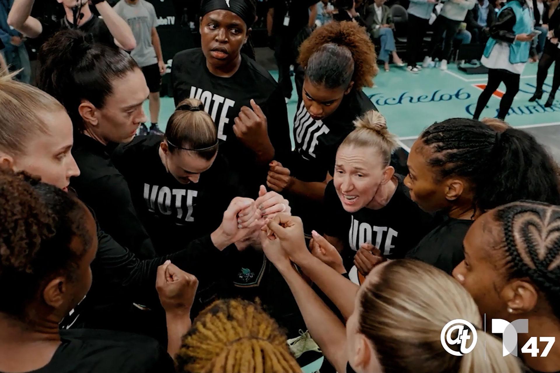A group of women athletes in black "VOTE" shirts stand in a huddle, hands joined in the center, showing unity and teamwork. The scene appears energetic and focused, with people watching in the background.