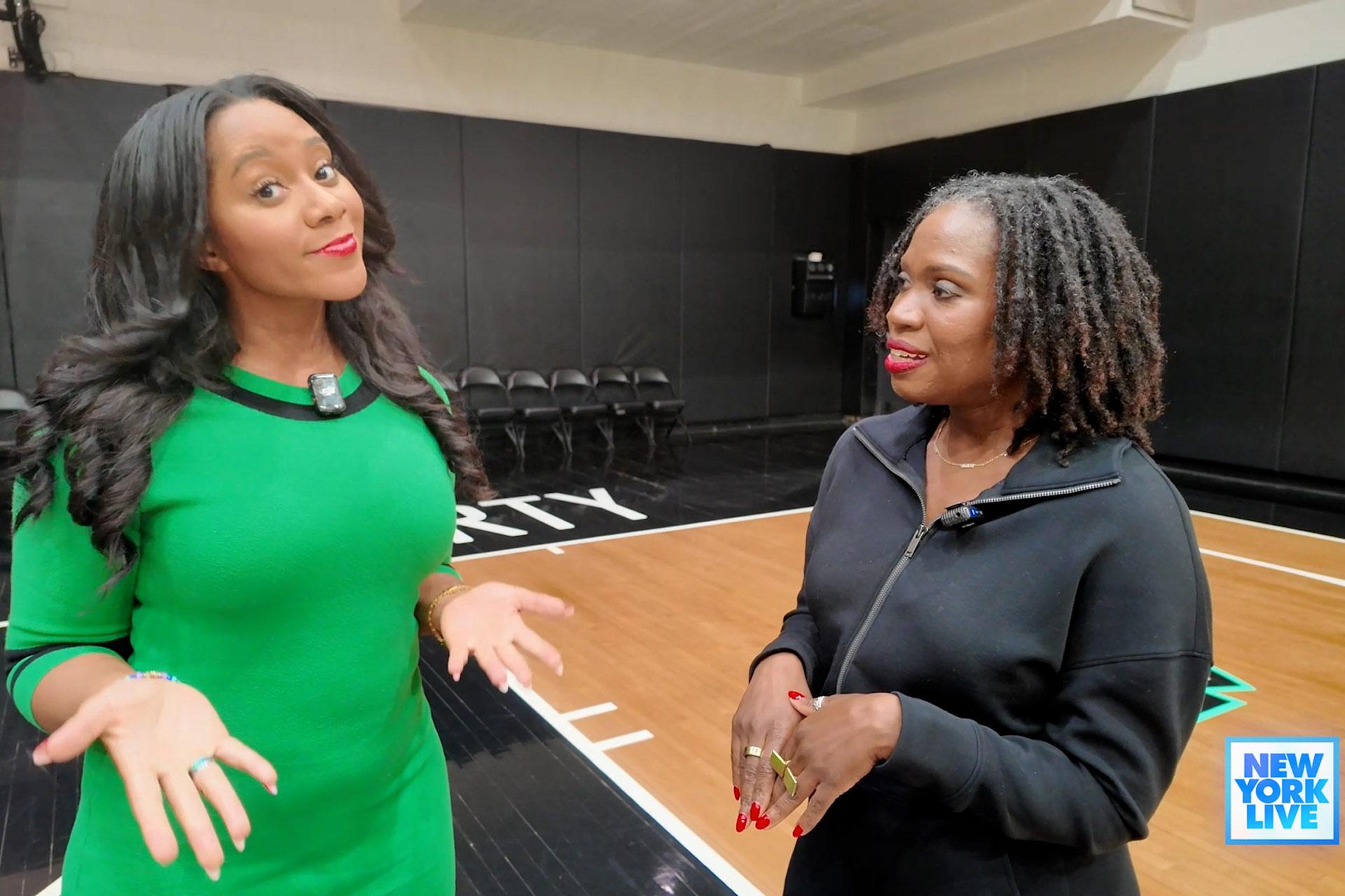Two women stand on an indoor basketball court, having a conversation. One wears a green dress and gestures with her hands, while the other, in a black outfit, listens attentively. The "New York Live" logo appears in the bottom right corner.