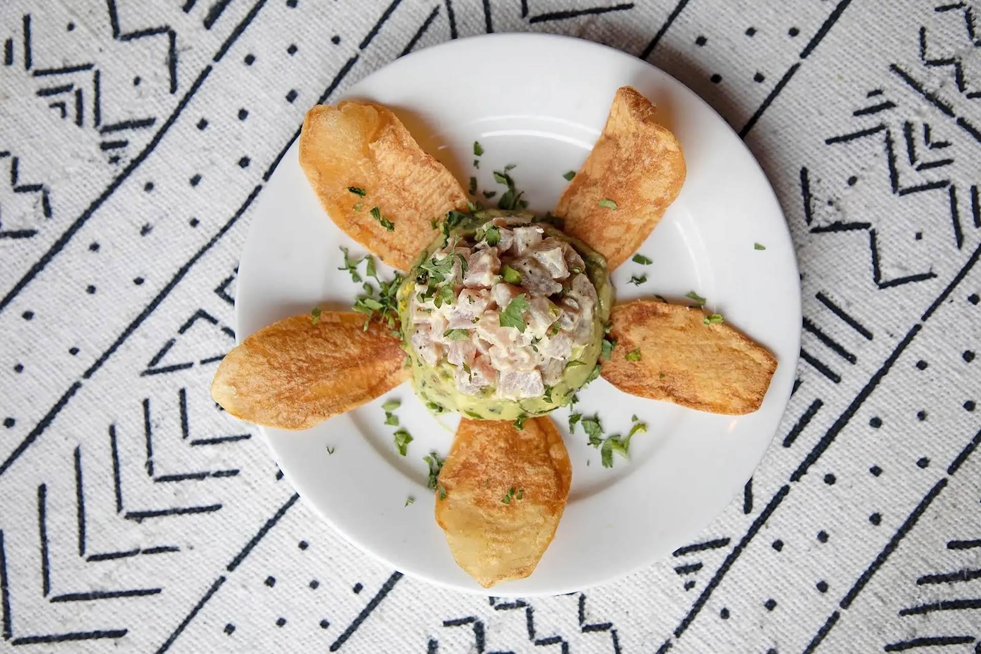 A white plate with a mound of tuna tartare garnished with herbs in the center, surrounded by five crispy potato chips arranged like petals. The plate is placed on a patterned black and white tablecloth.