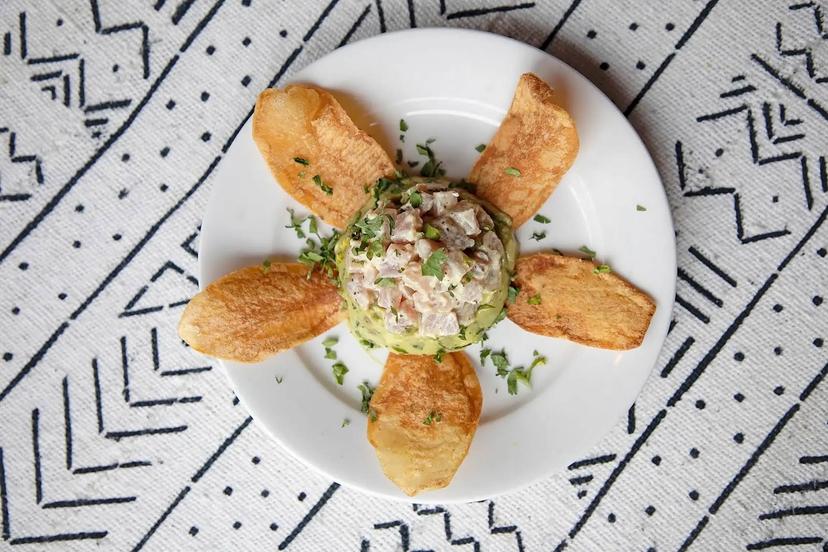 A white plate with a mound of tuna tartare garnished with herbs in the center, surrounded by five crispy potato chips arranged like petals. The plate is placed on a patterned black and white tablecloth.