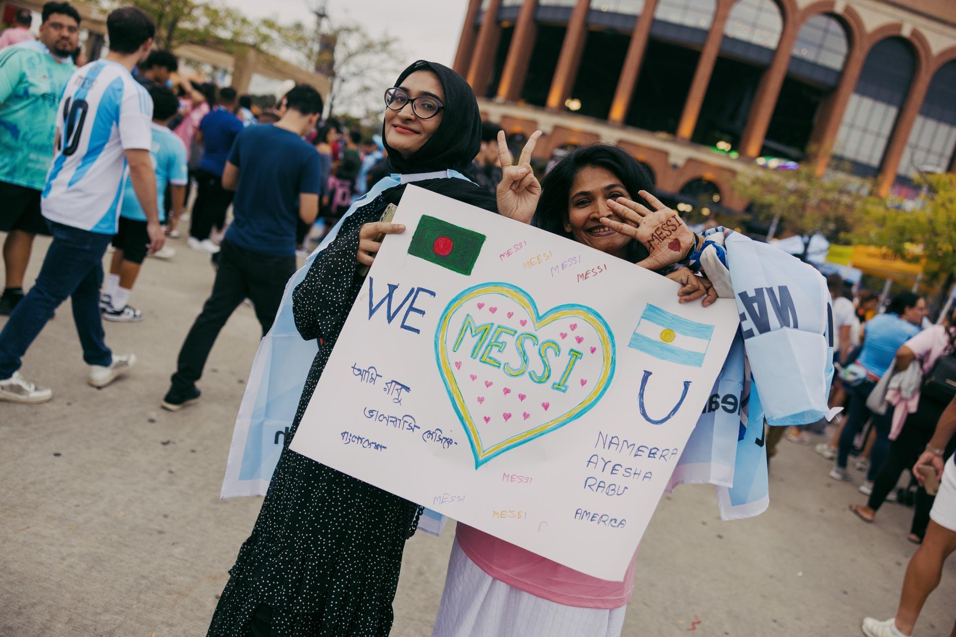 Two people smiling at the camera, one wearing a headscarf and holding a colorful "We Love Messi" sign at an outdoor event, surrounded by individuals in Argentina soccer jerseys. The sign is decorated with hearts, flags, and handwritten messages.
