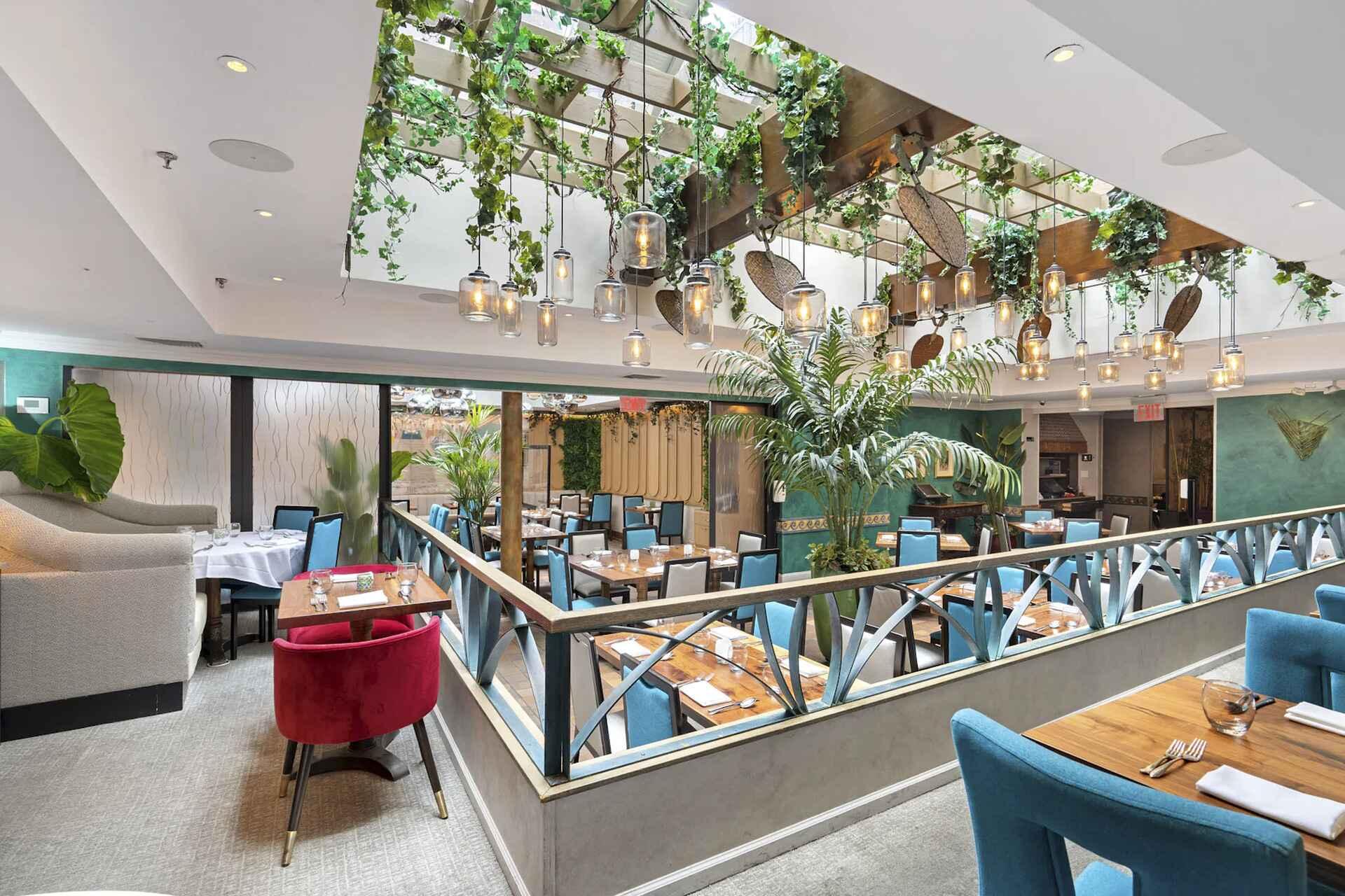 A modern restaurant with blue chairs, wooden tables, and a lush, plant-filled ceiling. Hanging lights and large windows provide natural light. There are green walls, a red armchair, and a relaxed, inviting atmosphere.