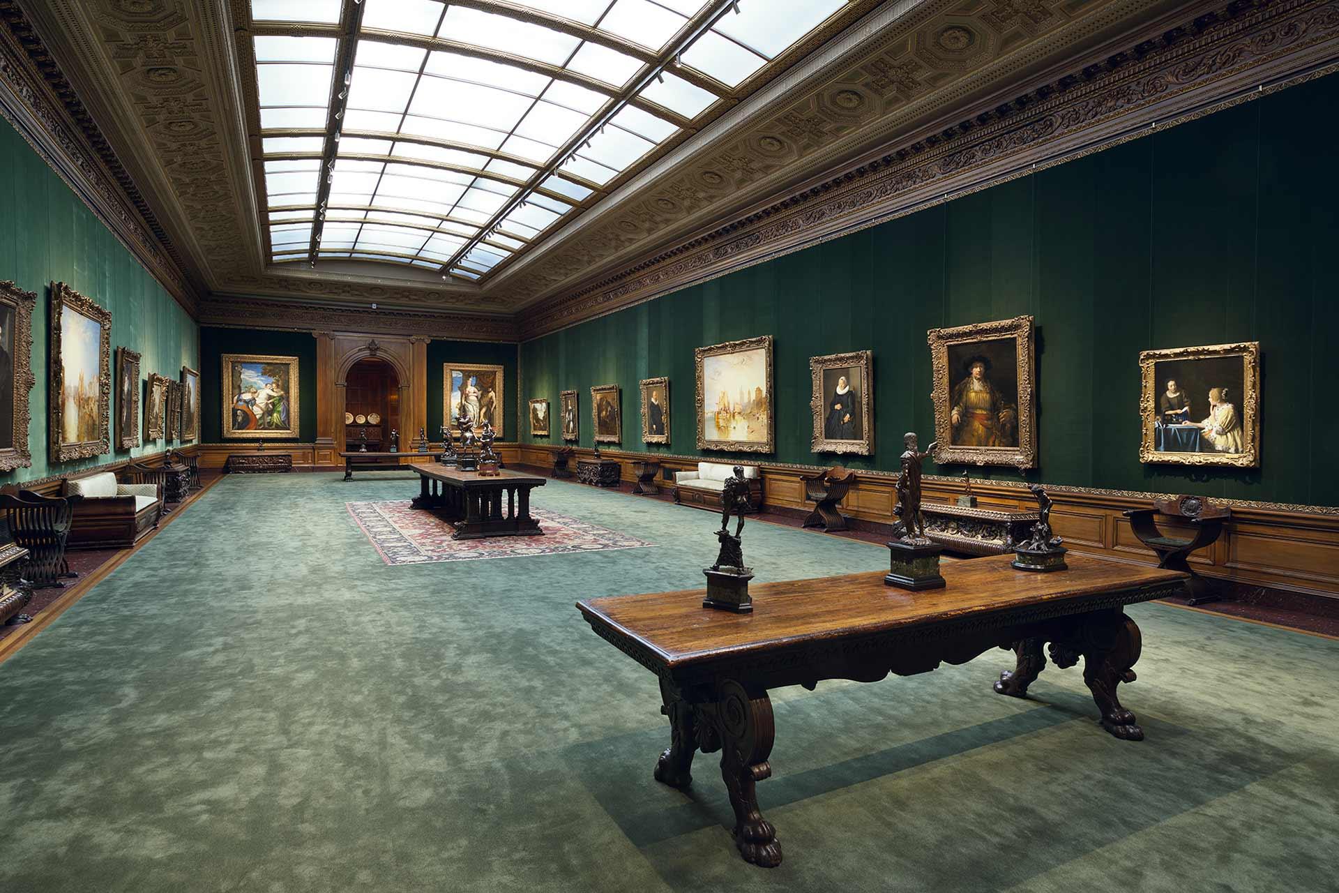 An elegant art gallery room with green walls, classic paintings in ornate frames, wood tables with small sculptures, a green carpet, and a vaulted glass ceiling letting in natural light.
