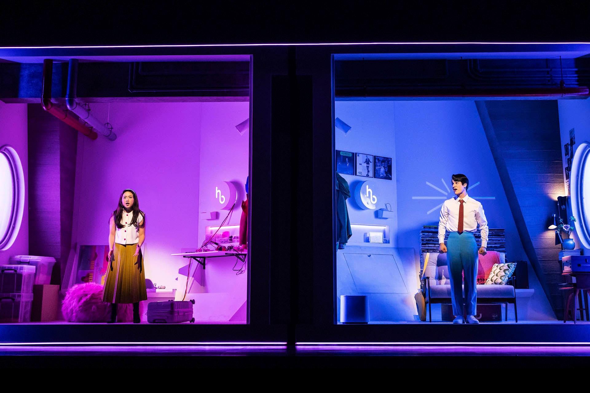 Two people in a split scene: the left side is lit in pink with a woman standing next to a bed, and the right side is lit in blue with a man standing near a desk. Circular neon lights enhance each side's color theme.