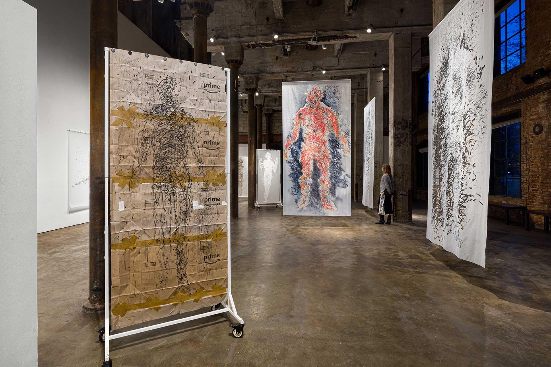 Art gallery with large abstract human figure artworks hanging from the ceiling; one piece is created on Amazon Prime packaging, others use various media. A person observes the art in the industrial-style space with concrete floors.