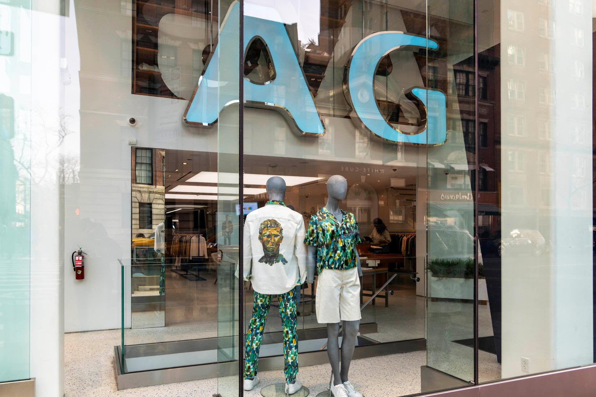 Storefront display with two mannequins dressed in colorful clothing, showcasing printed designs. Large blue "AG" letters are displayed above them. The store interior is visible through the glass window.