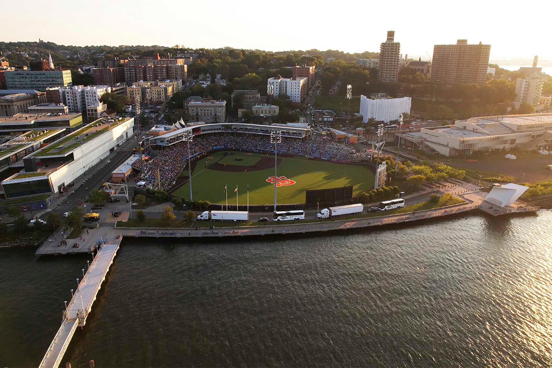 Aerial view of a riverside baseball stadium surrounded by urban buildings. The field is filled with spectators. A pier extends into the water, and the sun casts a warm glow over the scene.
