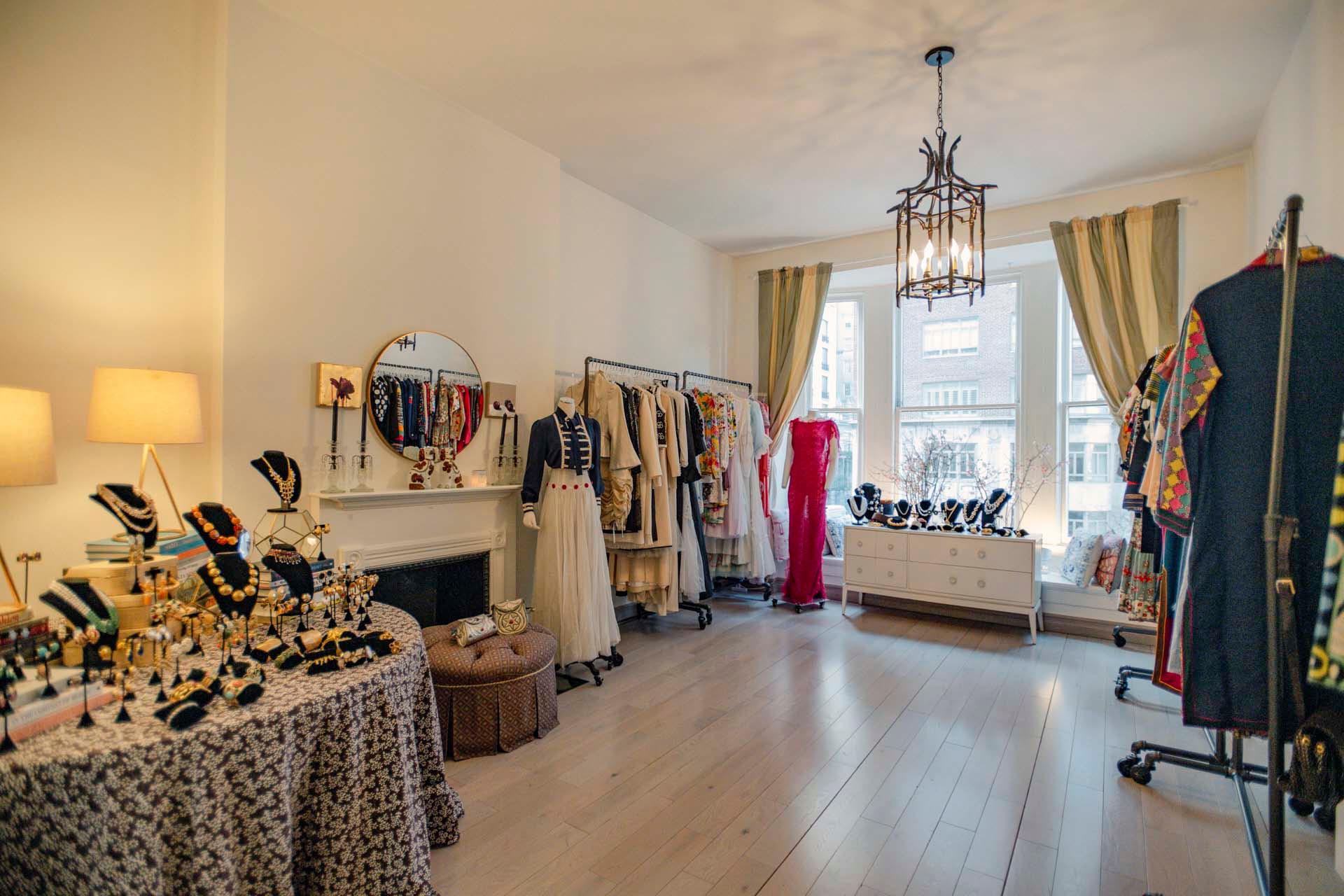 A boutique clothing store features elegant dresses on racks, a table with jewelry displays, and a cozy atmosphere with soft lighting. A large window with curtains illuminates the wooden floor, enhancing the chic and inviting setting.