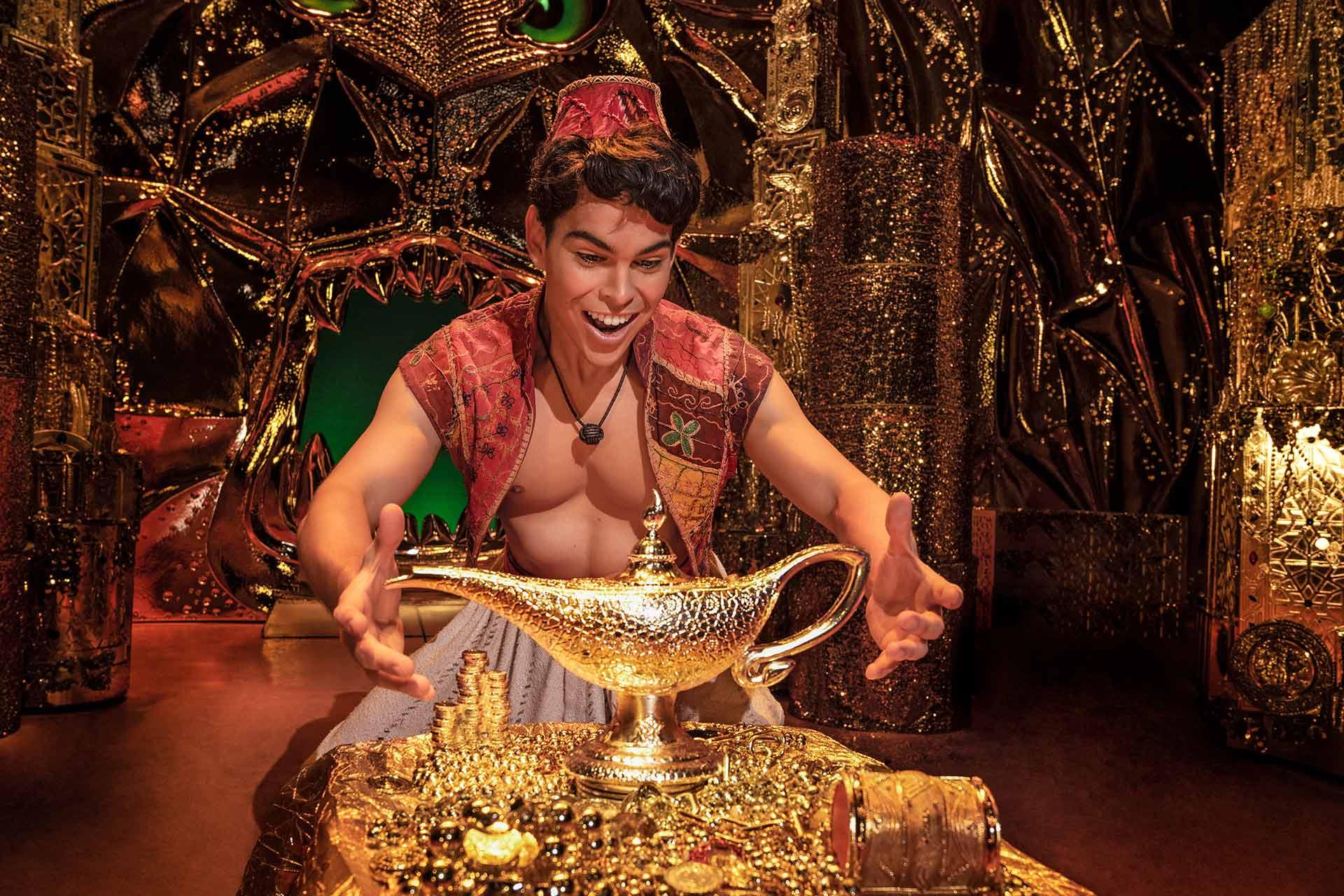 A young man dressed as Aladdin smiles excitedly with outstretched arms toward a large ornate magic lamp, surrounded by piles of gold and treasure in a richly decorated, golden cave.