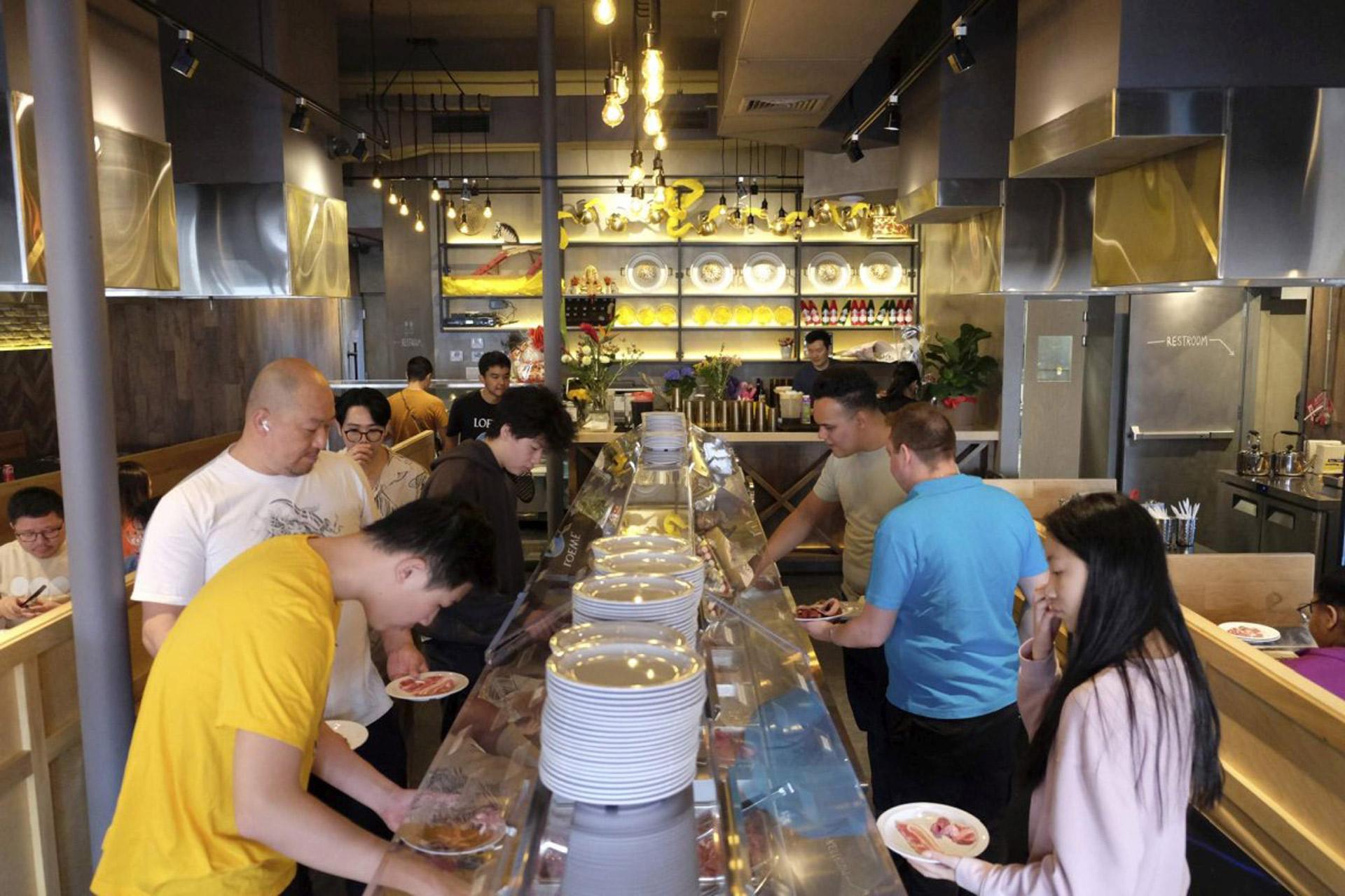 People are standing along a conveyor belt sushi bar, choosing plates of food. The restaurant features modern decor with warm lighting and shelves displaying various items. Diners are interacting and selecting dishes as the conveyor belt moves.