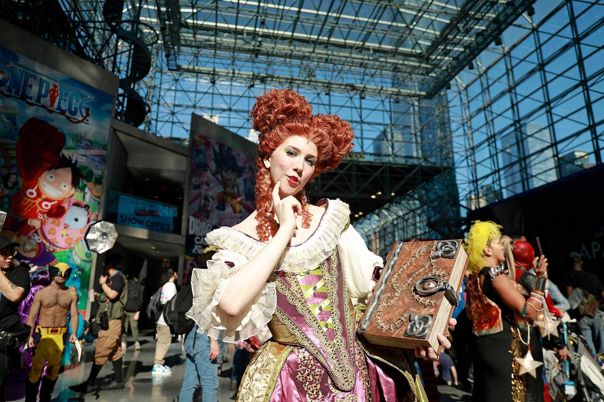 A person in an ornate red wig and elaborate historical costume holds a decorated book, posing at a lively convention center with cosplayers and colorful banners in the background.