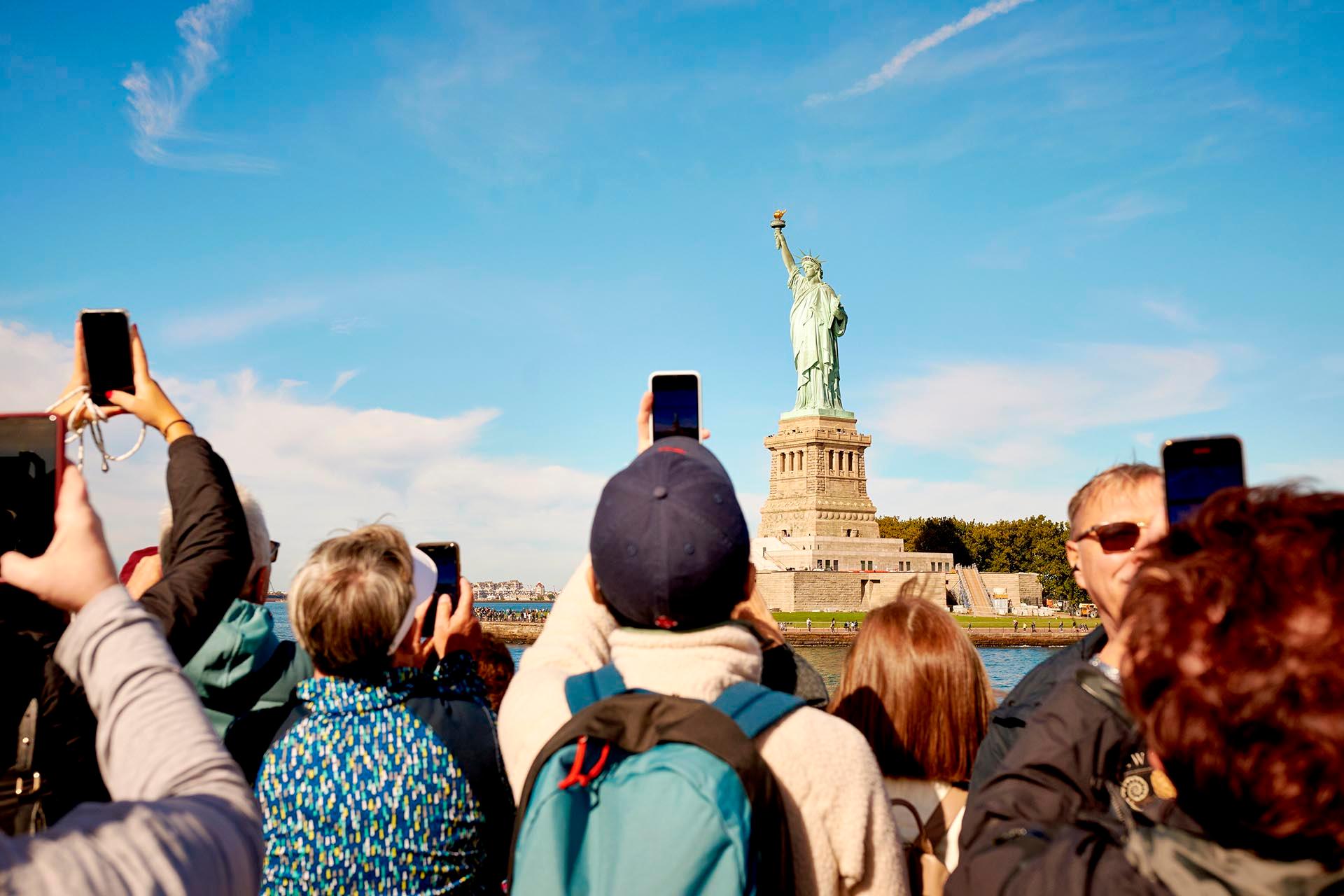 A group of people take photos with their phones of the Statue of Liberty on a sunny day, with blue sky and scattered clouds in the background.