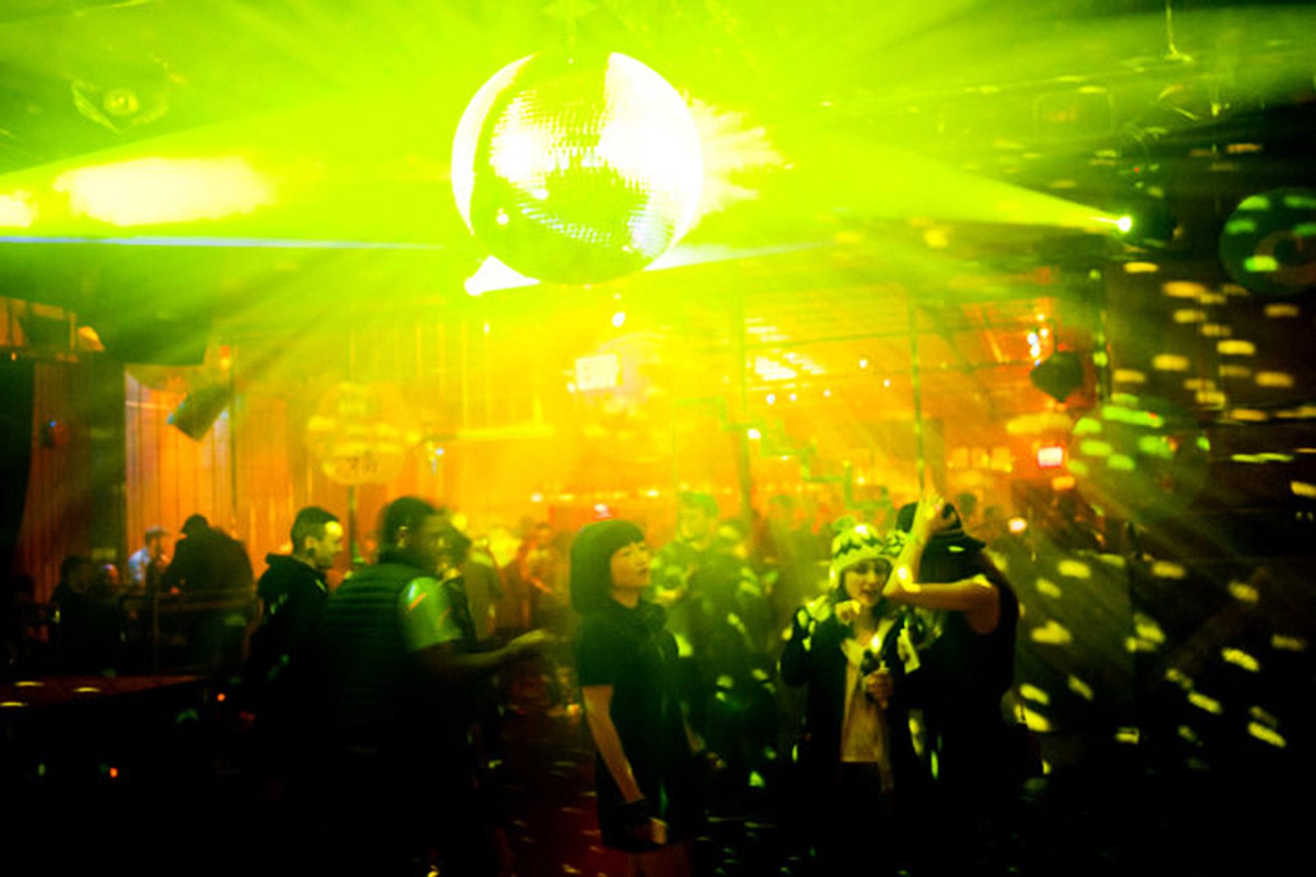 People dancing in a dimly lit nightclub with green and yellow lights shining from a disco ball, creating bright patterns across the room. The crowd enjoys the lively, energetic atmosphere.