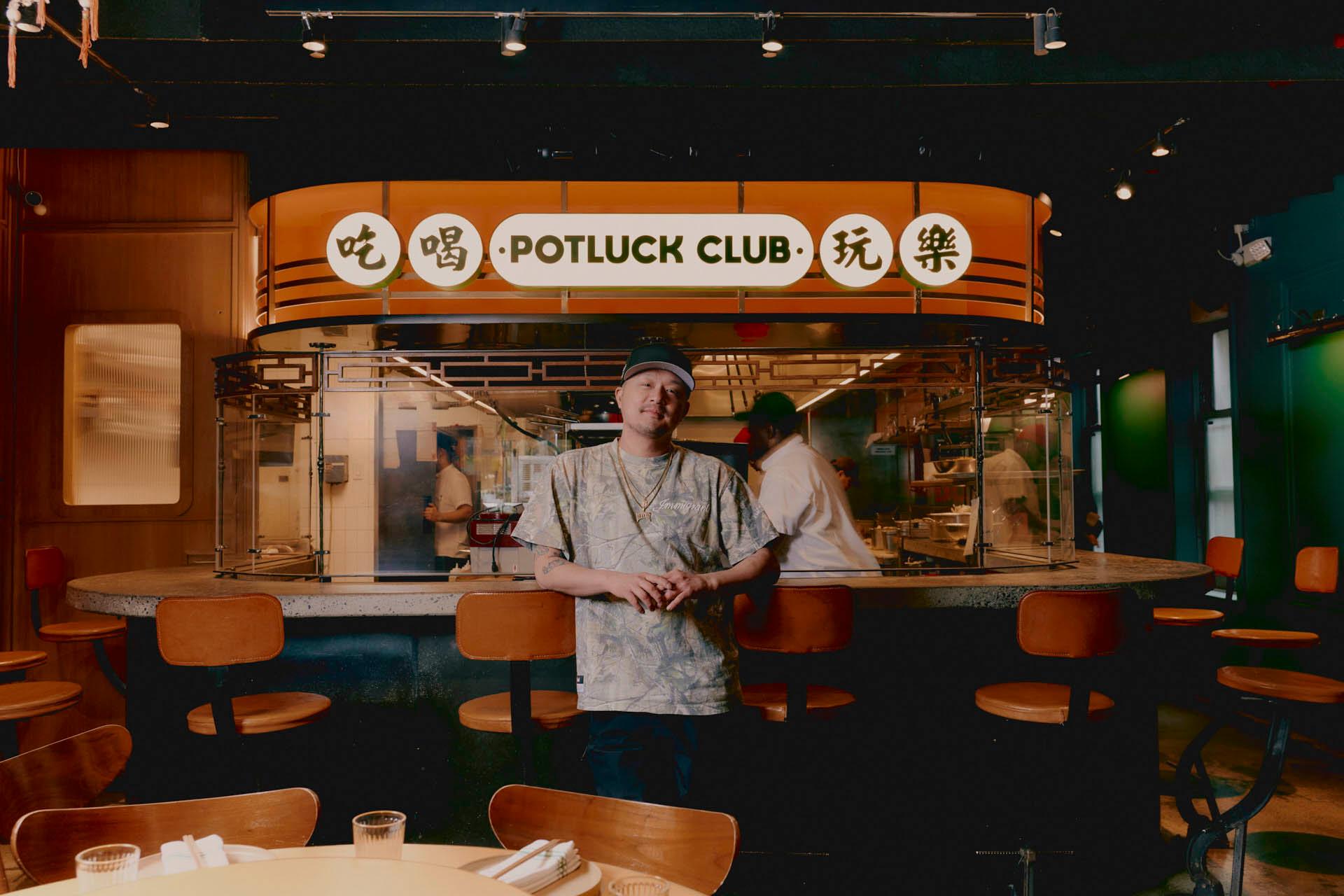 Cory Ng wearing a cap and casual clothes stands smiling in front of a restaurant counter with a "Potluck Club" sign in English and Chinese. The restaurant interior is modern, with chefs working in the background.