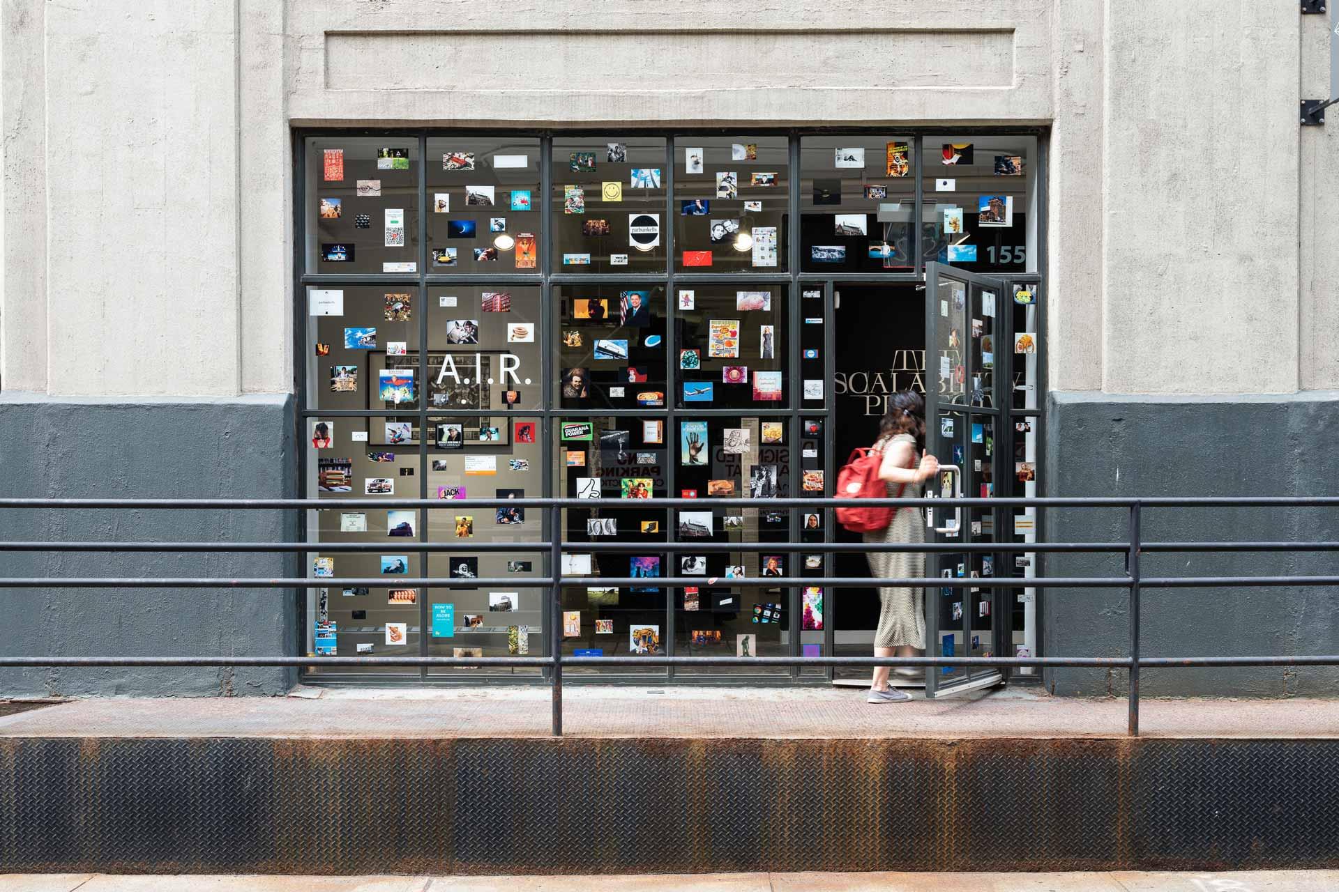 A person with a red backpack enters a gallery with large windows covered in many small, colorful photos. The gallery sign reads "A.I.R. SOA P P" and the building exterior is concrete with a black railing.