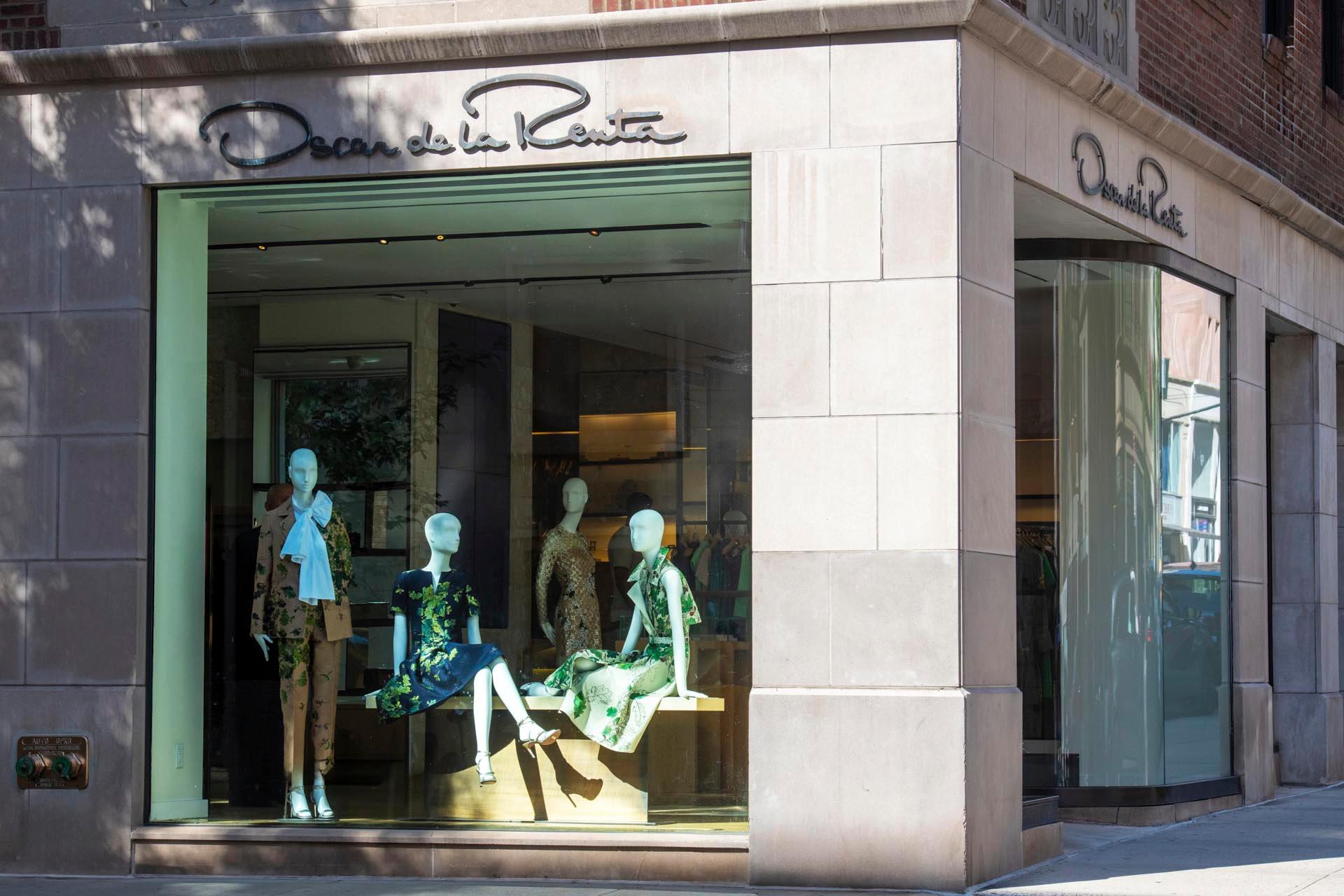 Storefront of Oscar de la Renta featuring mannequins displaying elegant dresses in green, blue, and floral patterns. The name is visible on the glass and building, with large windows showcasing the glamorous clothing inside.