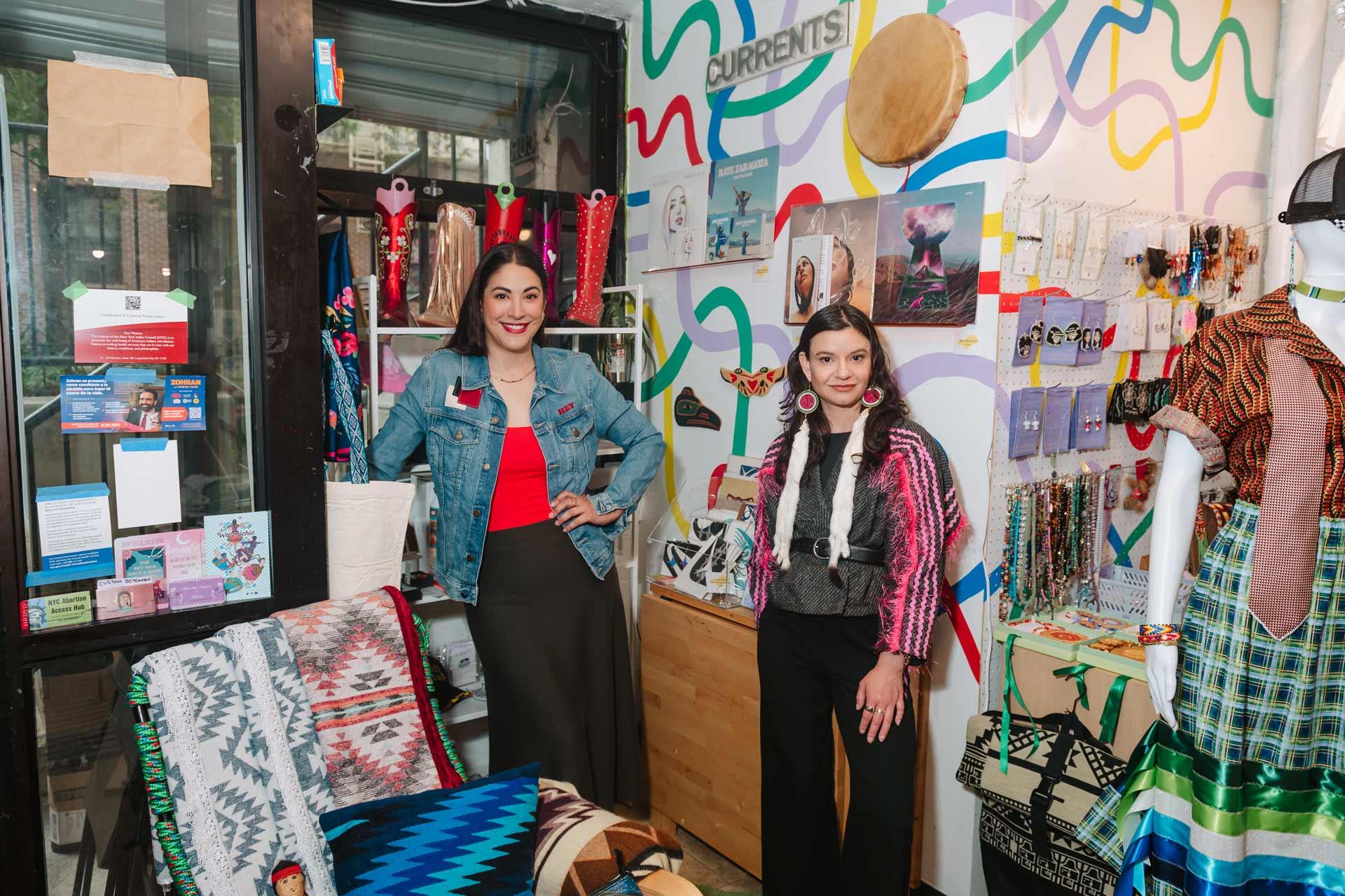 Two women stand inside a vibrant, colorful shop filled with textiles, jewelry, and Indigenous artwork. The walls are decorated with patterned art, and a mannequin displays traditional clothing.