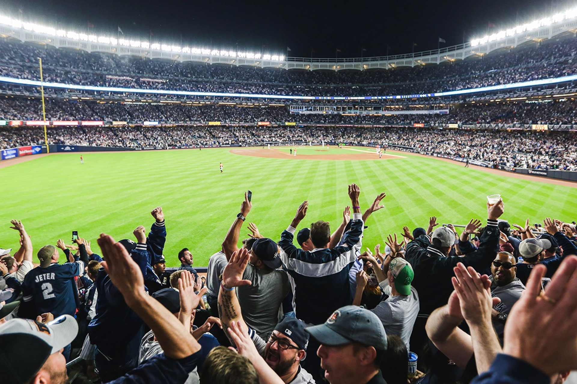 A large, enthusiastic crowd cheers in a brightly lit baseball stadium at night. Fans fill the stands, raising their arms toward the field, where players are visible near the infield under bright stadium lights.