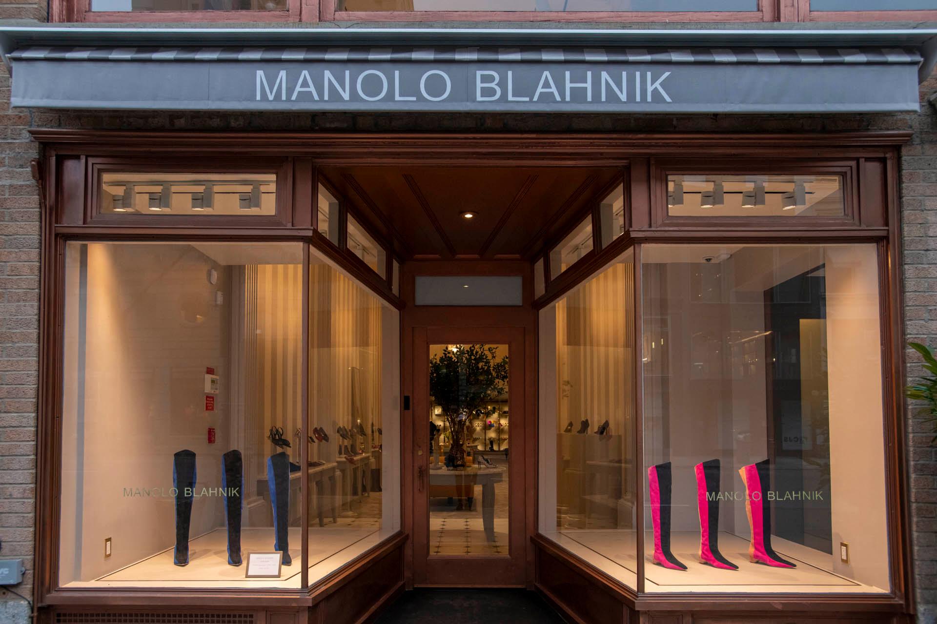 Storefront of a Manolo Blahnik shoe shop with large glass windows displaying various high-heeled boots. A dark wooden entrance is centered between the window displays. The shop's name is on a gray sign above the entrance.