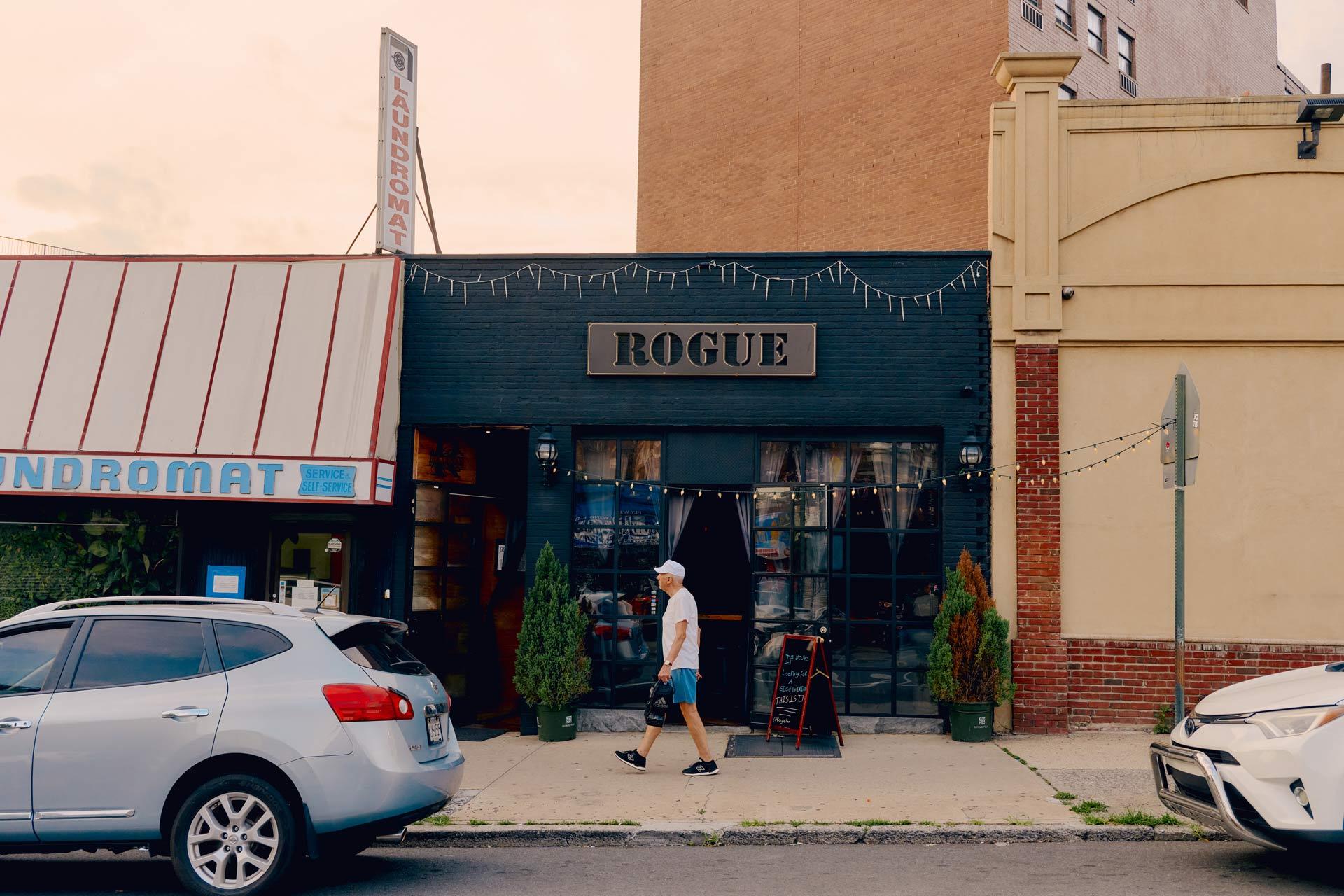 A man walks past parked cars and a bar called "ROGUE" with string lights above the entrance. The bar is on a city street, next to a laundromat and another building.