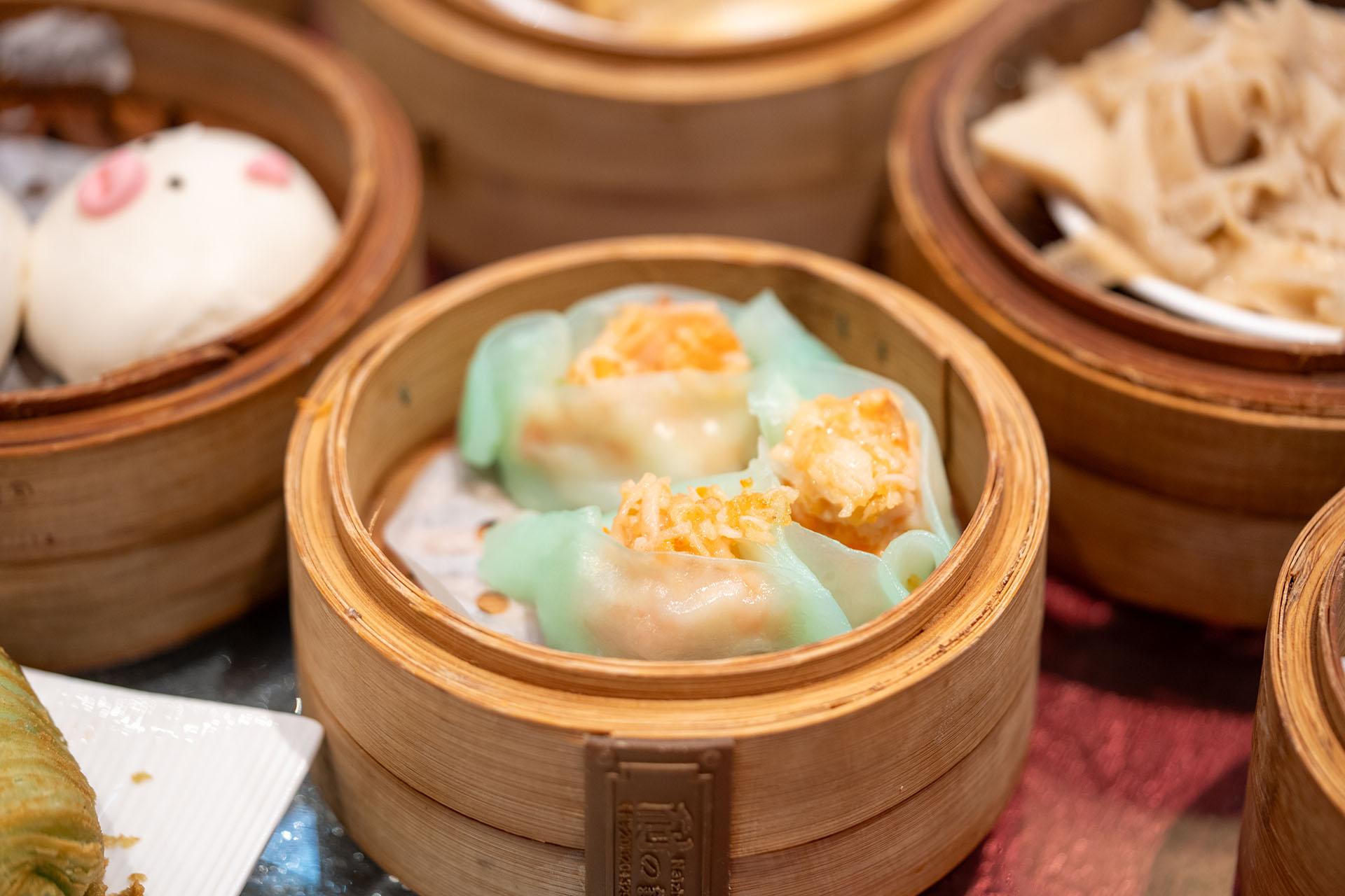 A bamboo steamer filled with two shrimp dumplings wrapped in translucent greenish wrappers, surrounded by other dim sum dishes on a table.