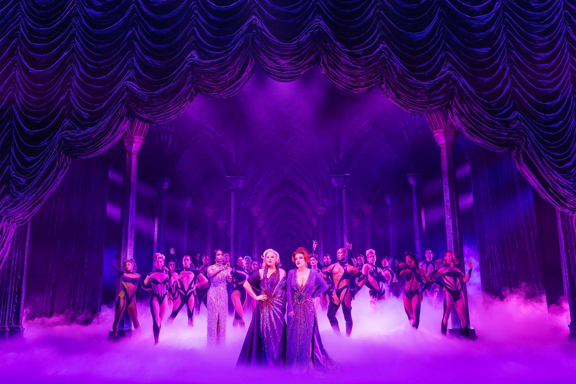 A dramatic theatre stage with purple lighting, featuring two performers in sparkling gowns at the front, surrounded by a group of dancers in matching costumes. The background displays elegant, draped curtains and columns