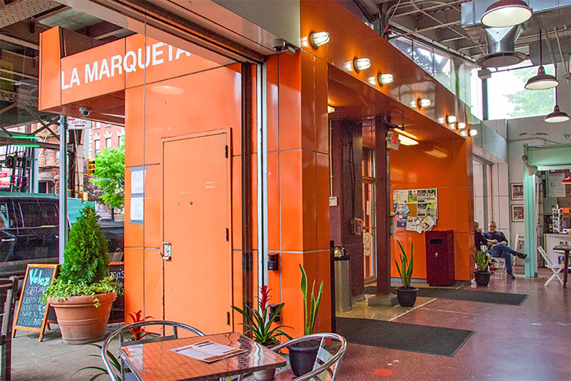 The entrance to La Marqueta features orange walls, potted plants, a chalkboard menu, and a small table with chairs. Inside, a man sits near a bulletin board under an industrial-style ceiling.