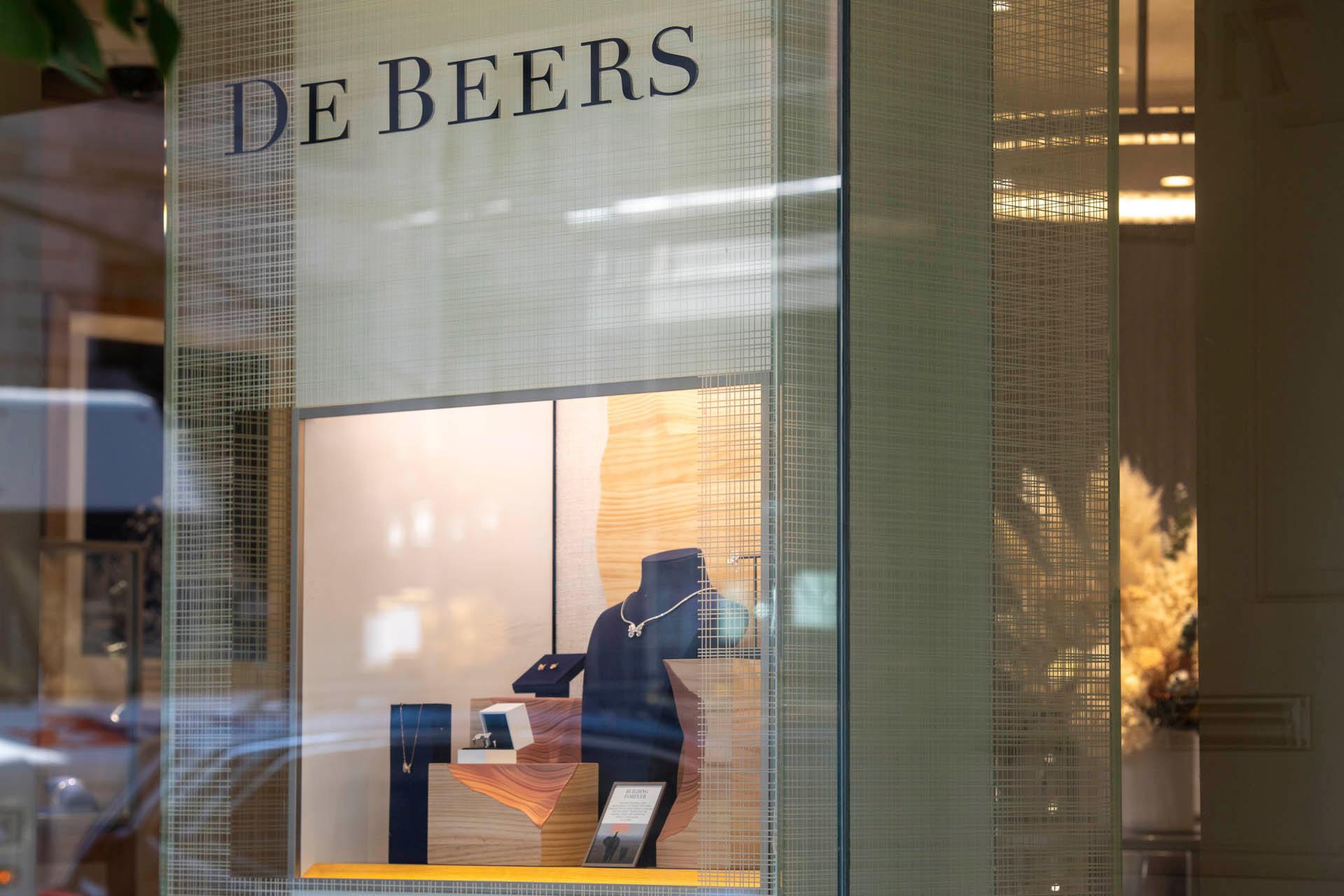 Jewelry display in a store window with the "De Beers" sign above. The display features necklaces and rings on stands, with soft lighting and a textured background, viewed through glass with reflections.