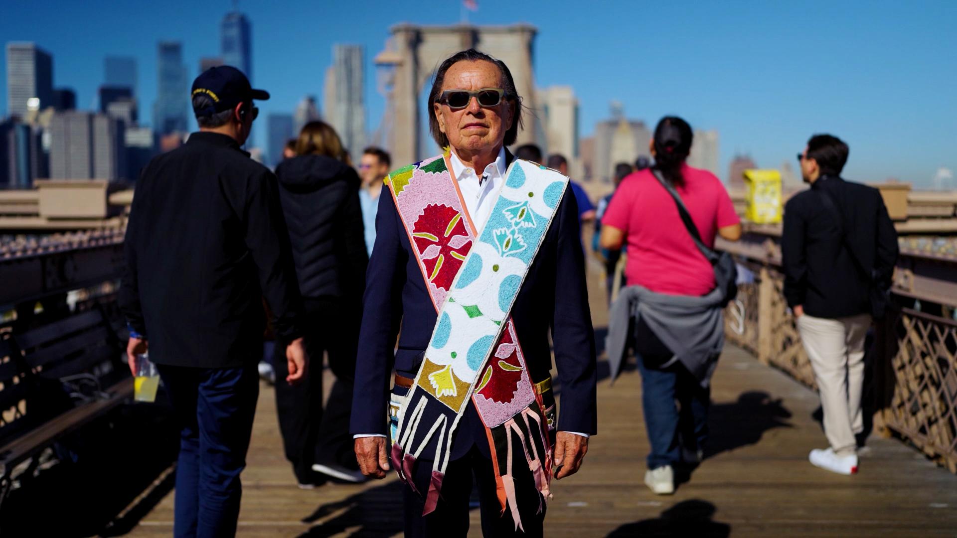 A person wearing sunglasses and a colorful patterned scarf over a suit stands on a bridge with a clear, sunny sky. City buildings are visible in the background. Other people are walking on the bridge.