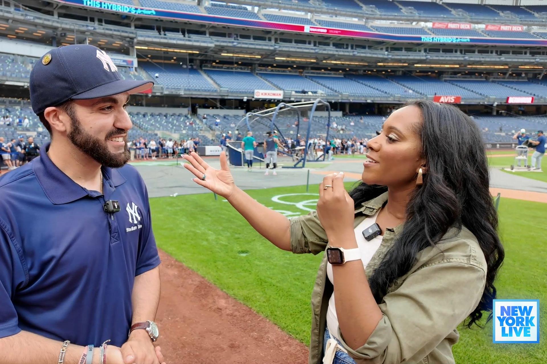 A woman interviews a man in a New York Yankees polo shirt on a baseball field. They are smiling and gesturing as they talk, with the stadium seats and a batting cage visible in the background.