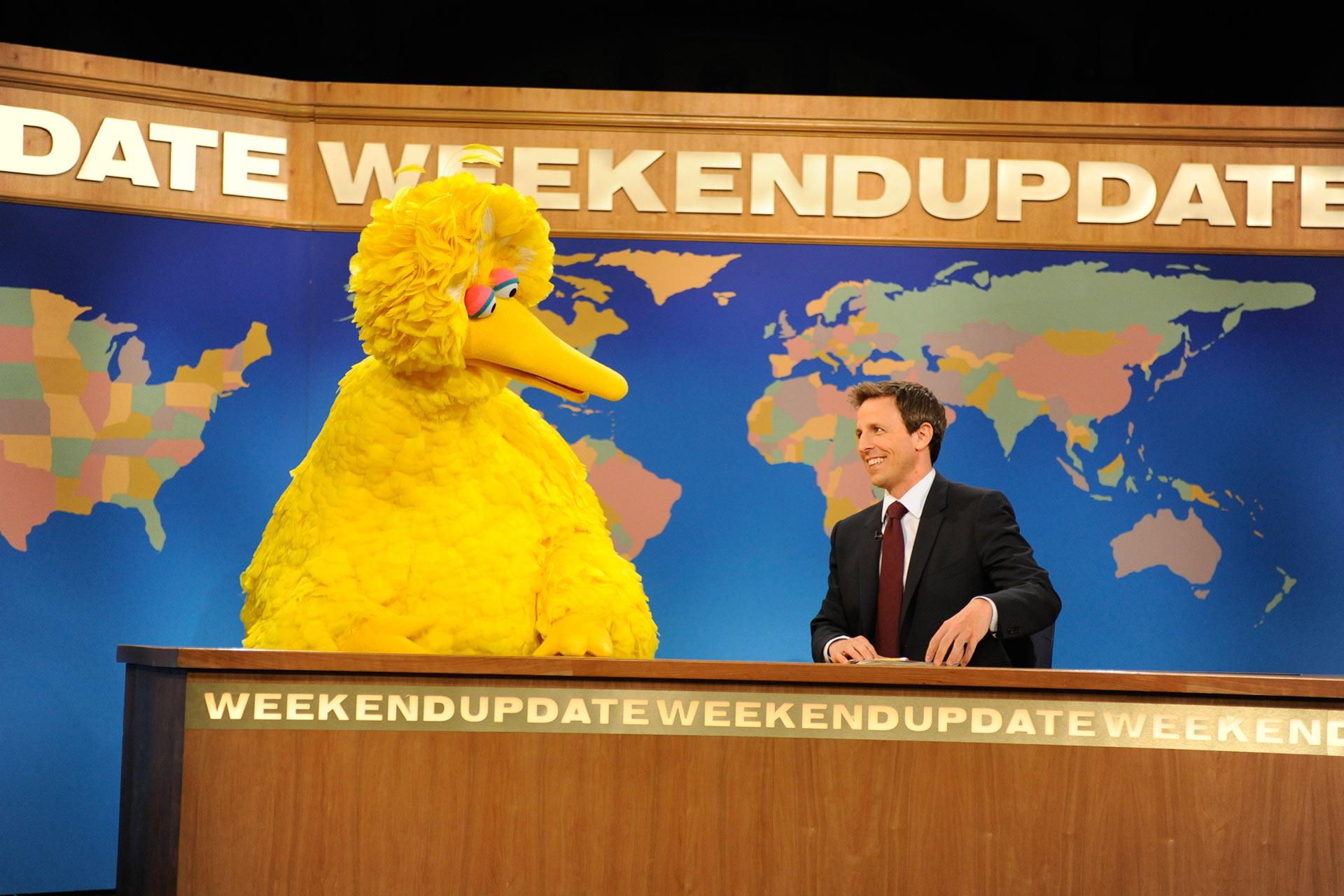 Big Bird sits at a news desk next to a smiling man in a suit. The backdrop shows a world map and the words "WEEKEND UPDATE." Both appear to be engaged in a lighthearted conversation.