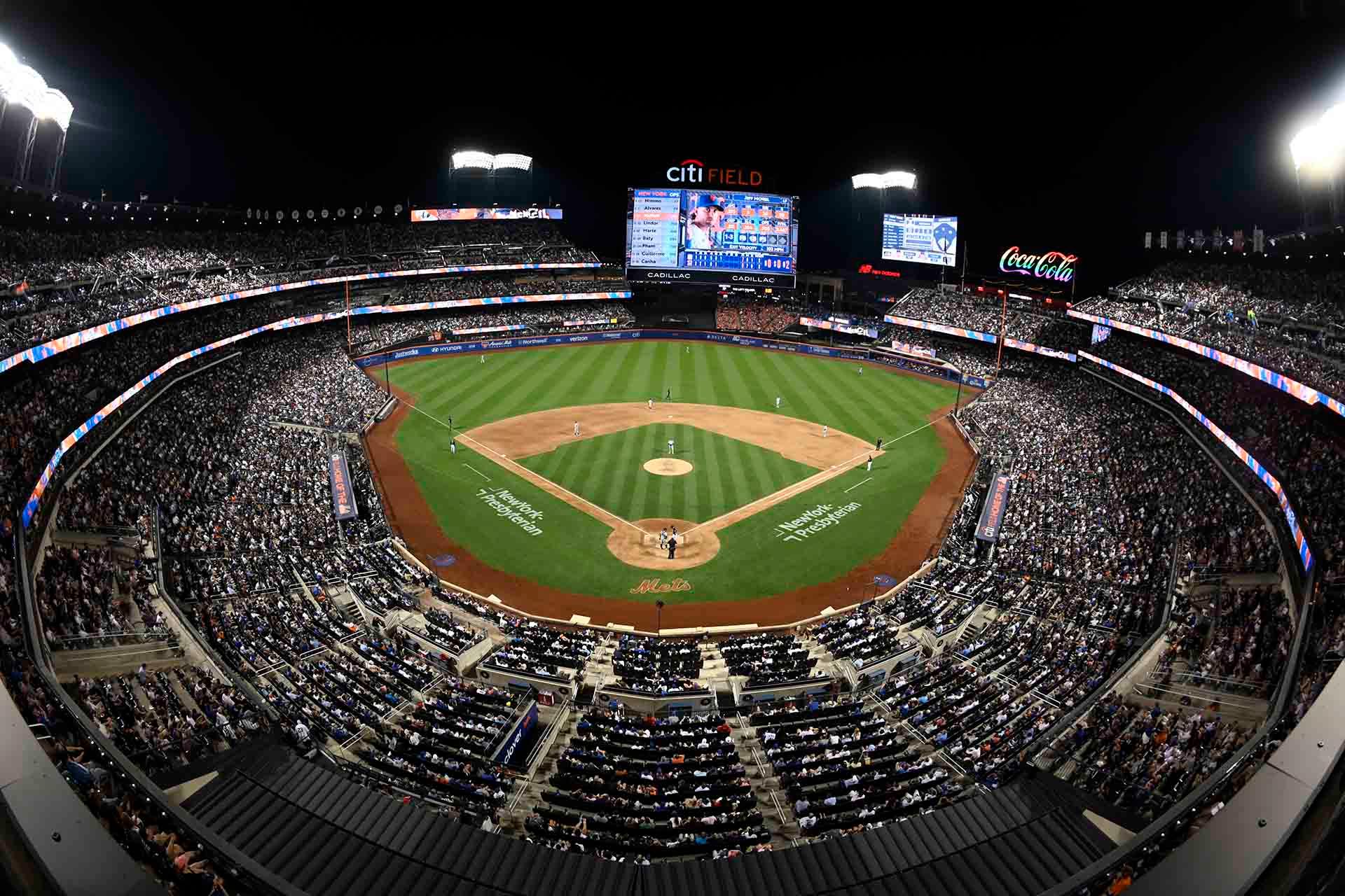 A nighttime, wide-angle view of a packed baseball stadium during a game, with bright lights illuminating the field and large crowds filling the stands. The scoreboard and advertisements are visible in the background.
